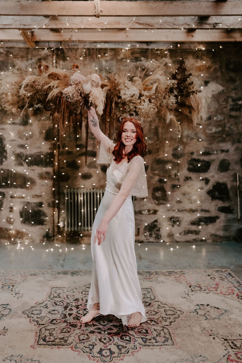 Bride is stood barefoot holding up her bridal bouquet. She is wearing a long cream sleek wedding dress with a sheer gold beaded layer over the bust and flowing into soft batwing sleeves.