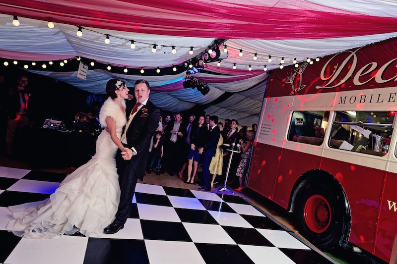  Bride and groom are dancing on the black &amp; white dance floor which is placed outside, under a canopy of red and white fabric which are the same colours as the vintage bus it is next to.  Photographer Credit: Adrian Jones Photography 