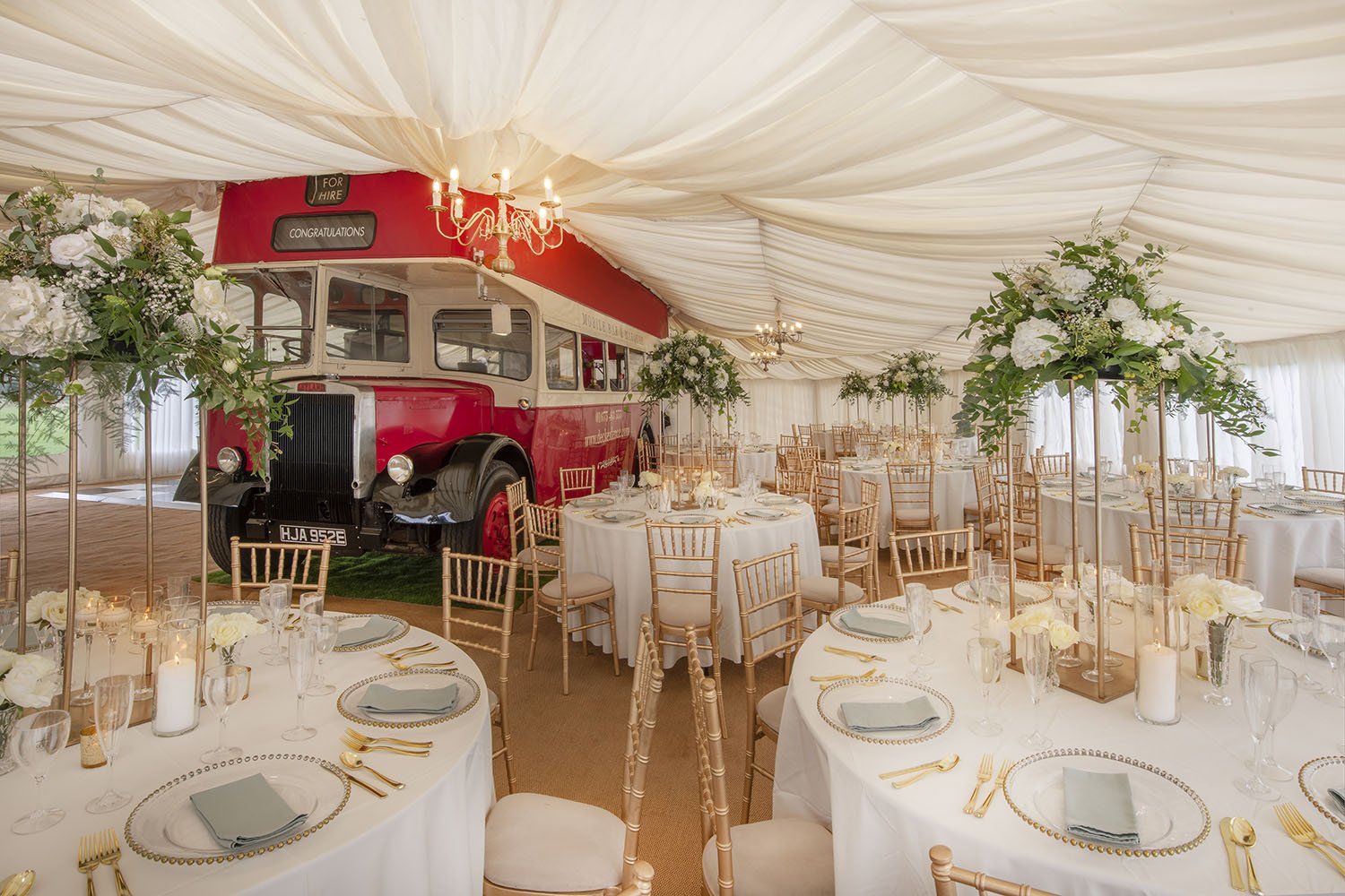  The vintage bus is a unique wedding venue and is a prominent feature surrounded by chairs and round tables laid for a wedding breakfast. There is cream fabric draped above to hide the ceiling of the marquee whilst the addition of wedding florals and