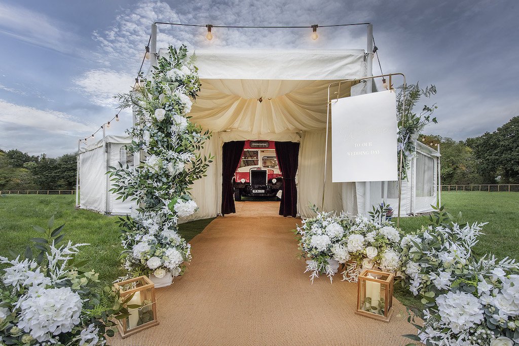  The front of the retro bus can be seen through the opening of a marquee at the end of an aisle lined with white wedding florals and storm lanterns with pillar candles. Lightbulbs are attached to the suspended metal frame outside. 