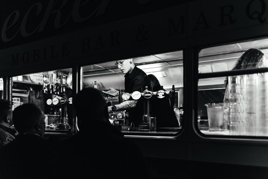  A black and white image of a bartender serving wedding guests through the windows of the retro bus which is a unique mobile wedding venue.  Photographer Credit: Electric Blue Photography 