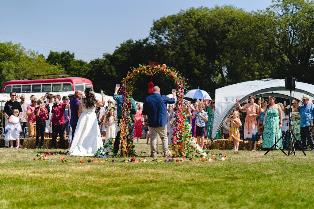  The photographer is stood behind the bride and groom who can be seen greeting their wedding guests outside on a bright sunny day. A floral arch is to the left of the image, whilst the top of the double decker, vintage red and cream bus can be seen t