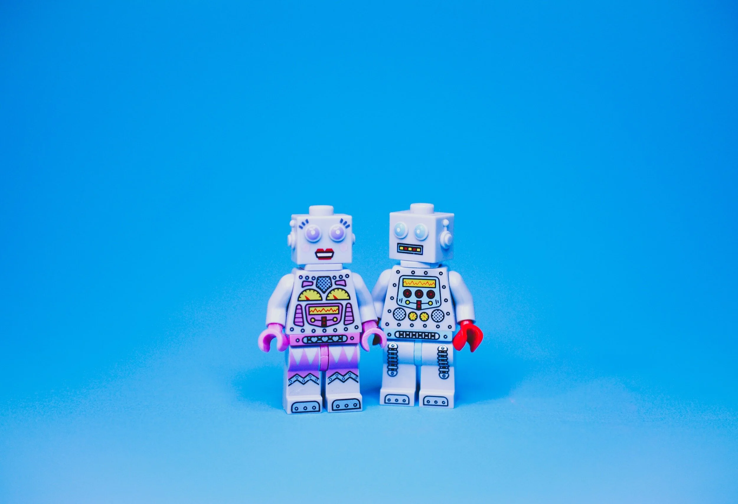 Two lego robots stood next to each other in front of a bright blue background.