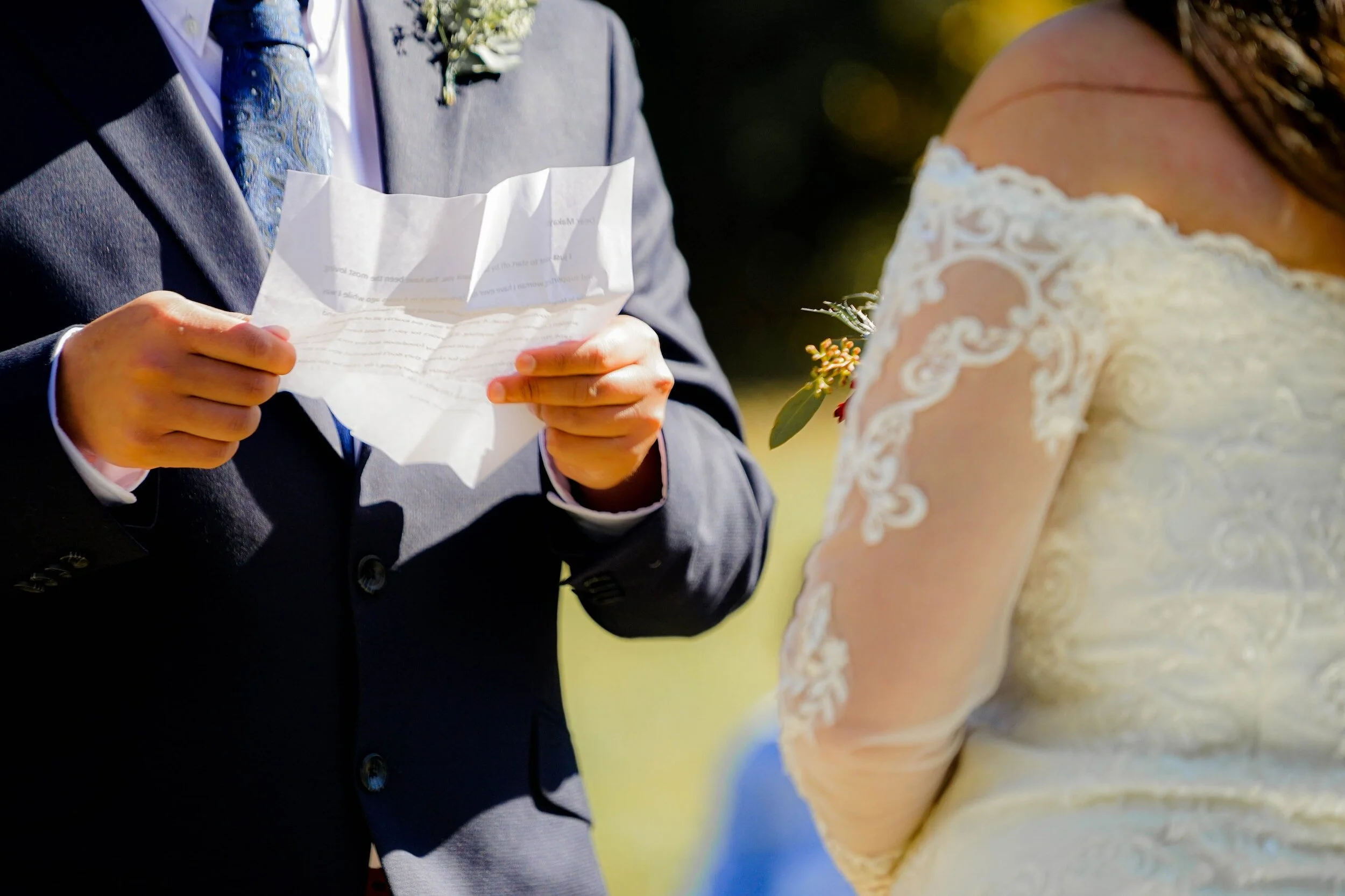 A groom holding his wedding vows which are printed on a piece of paper. The bride is wearing a long sleeved white wedding dress and the groom is wearing a navy suit, white shirt and bright blue tie.