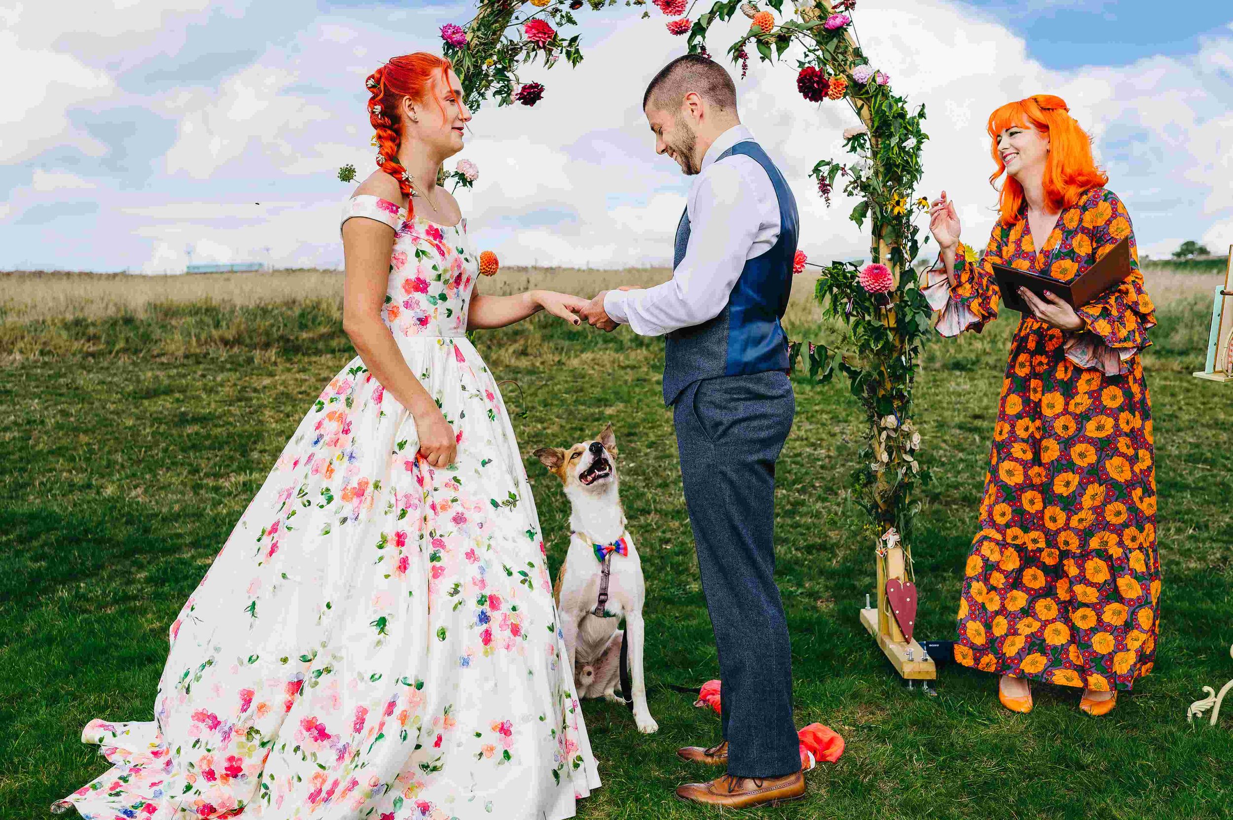  Photo by: Andrew Craner  A bright image of a cool humanist wedding celebrant standing to the side of the bride and groom in front of a floral arch. She is wearing a bright, long sleeved, long dress covered in a bright orange flower print with orange