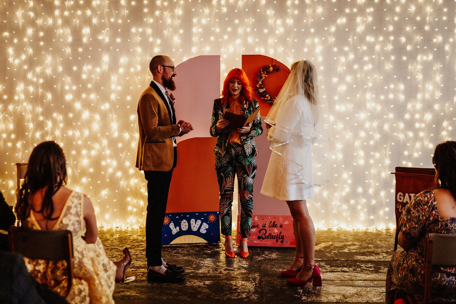  Photo by: Andrew Heeley Photography  Image shows humanist celebrant in front of a wall of fairy lights and a bright ceremony backdrop in orange and pink with LOVE IS LIKE A BUTTERFLY printed on.  The wedding celebrant is wearing a bright palm print 