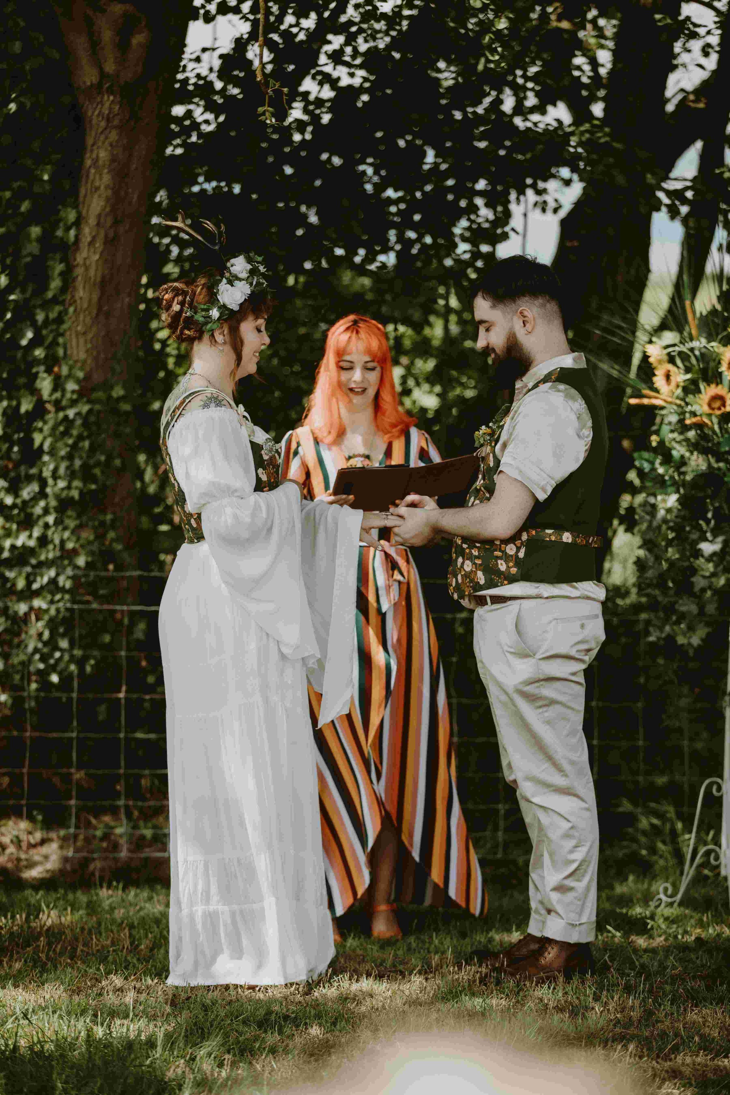  Photo by: Christopher Terry Photography  Wearing a stripy, short sleeved wrap dress the cool, modern wedding celebrant is stood reading from her folder, whilst the bride and groom are stood in front of her facing each other holding hands. The humani