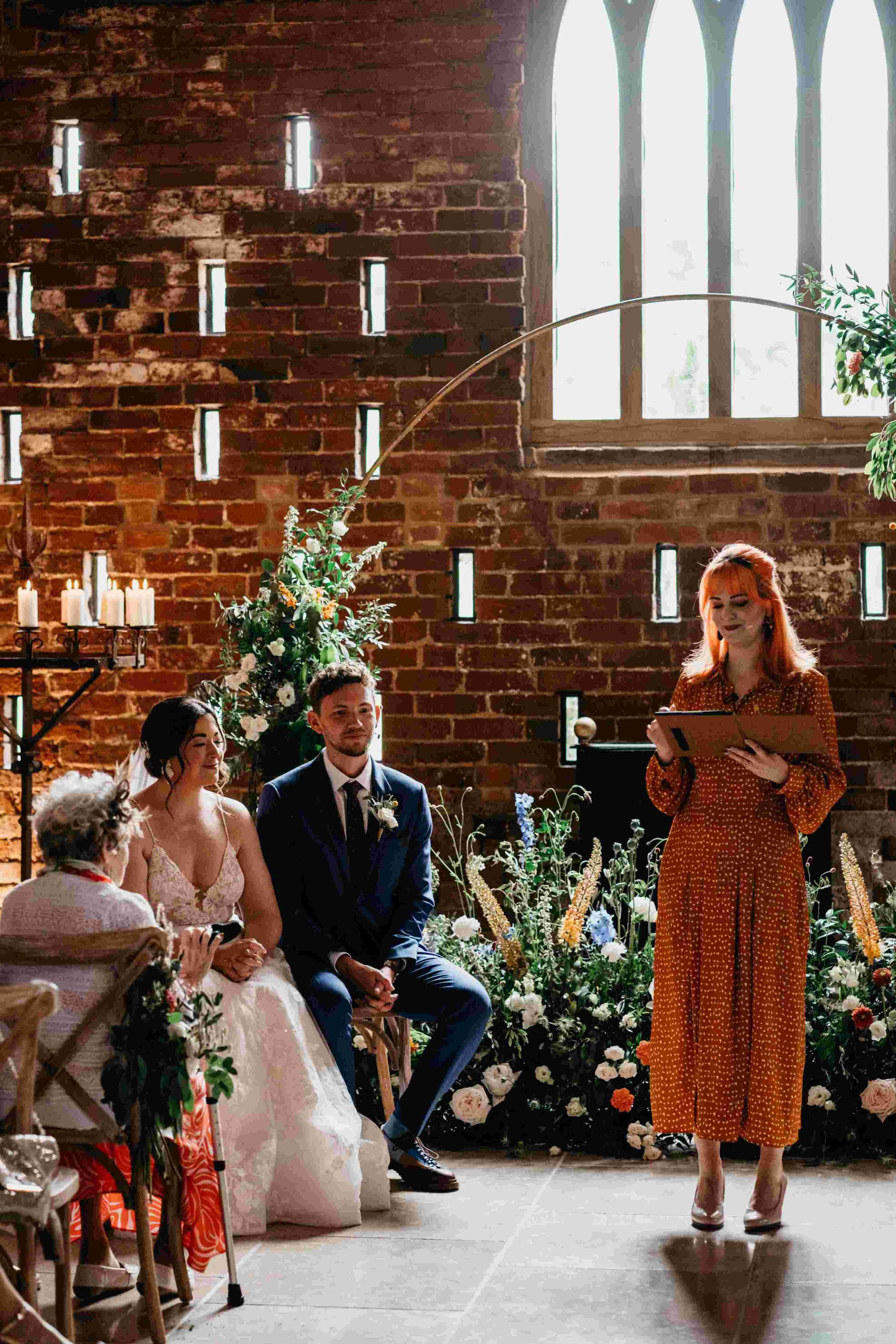  Photo by: David Boynton Photography  The modern celebrant is stood in a beautiful room with exposed brick walls and grand arched stone windows as well as lots of brick sized, small rectangle windows allowing lots of light to flood in.   There is a l