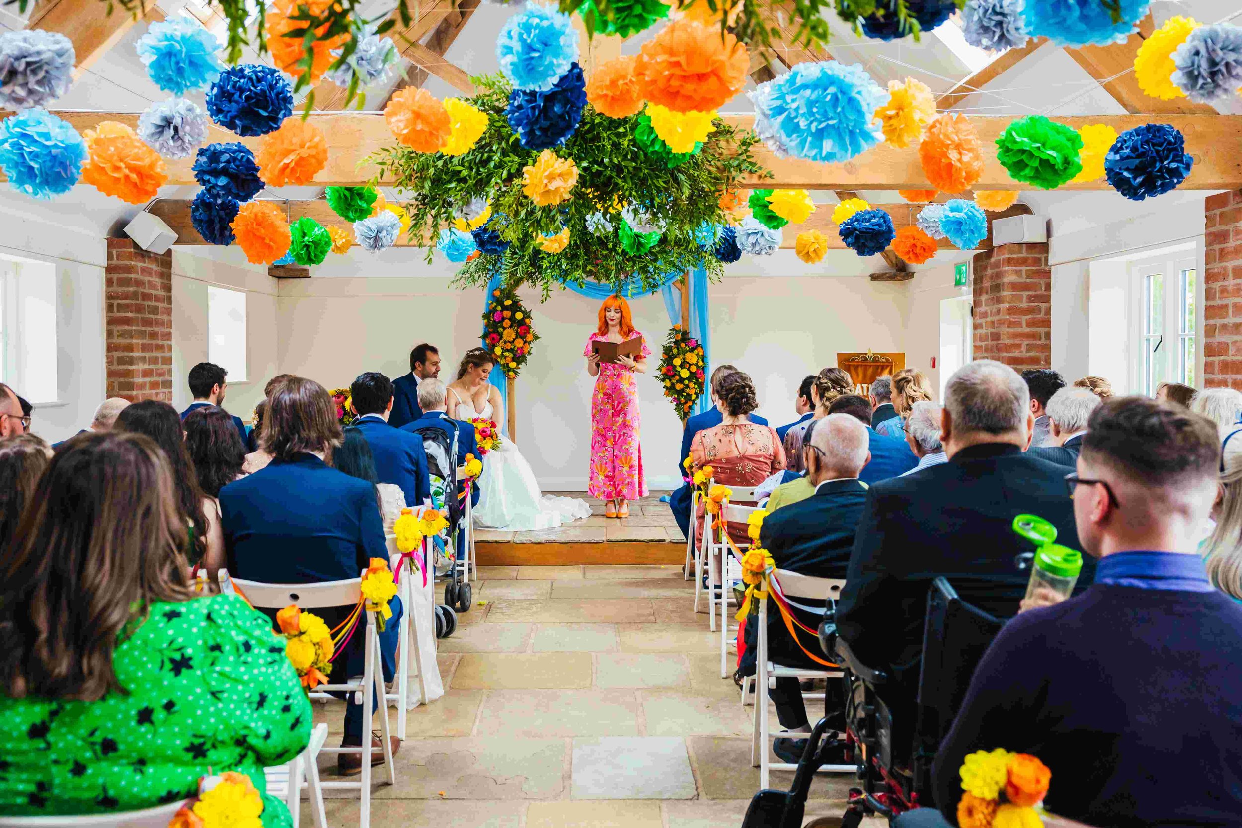  Photo by: Kirsty Rockett Photography    Vibrant image of a modern humanist celebrant conducting the wedding ceremony. The wedding celebrant is stood facing the wedding guests with the bride and groom sat to the side of her.   From the bleached woode