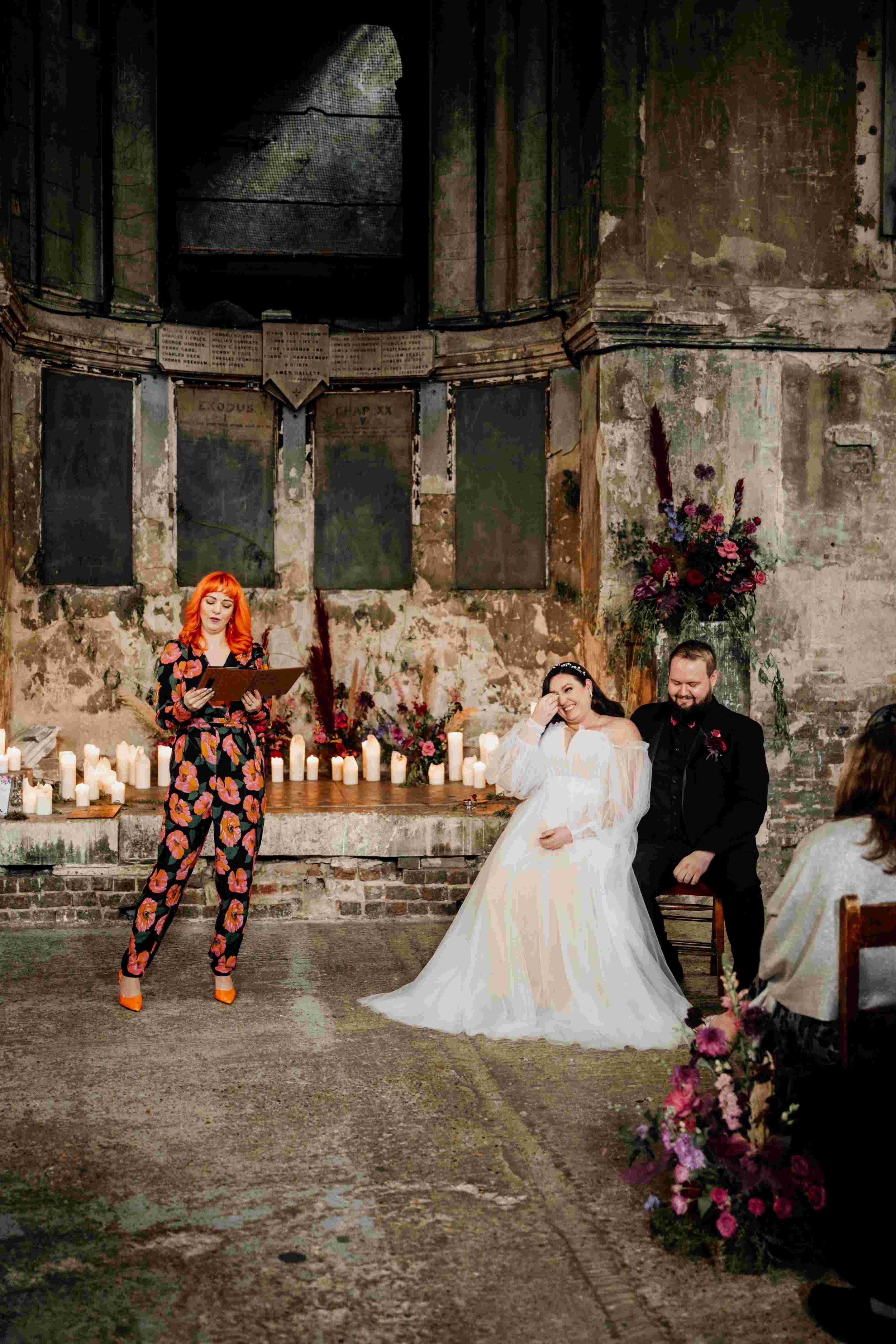  Photo by: Tom Wyatt Photography  Within a bare stone room and with a low ledge filled with pillar candles and a wild floral installation which the modern wedding celebrant is stood in front of facing the bride and groom’s wedding guests with the bri