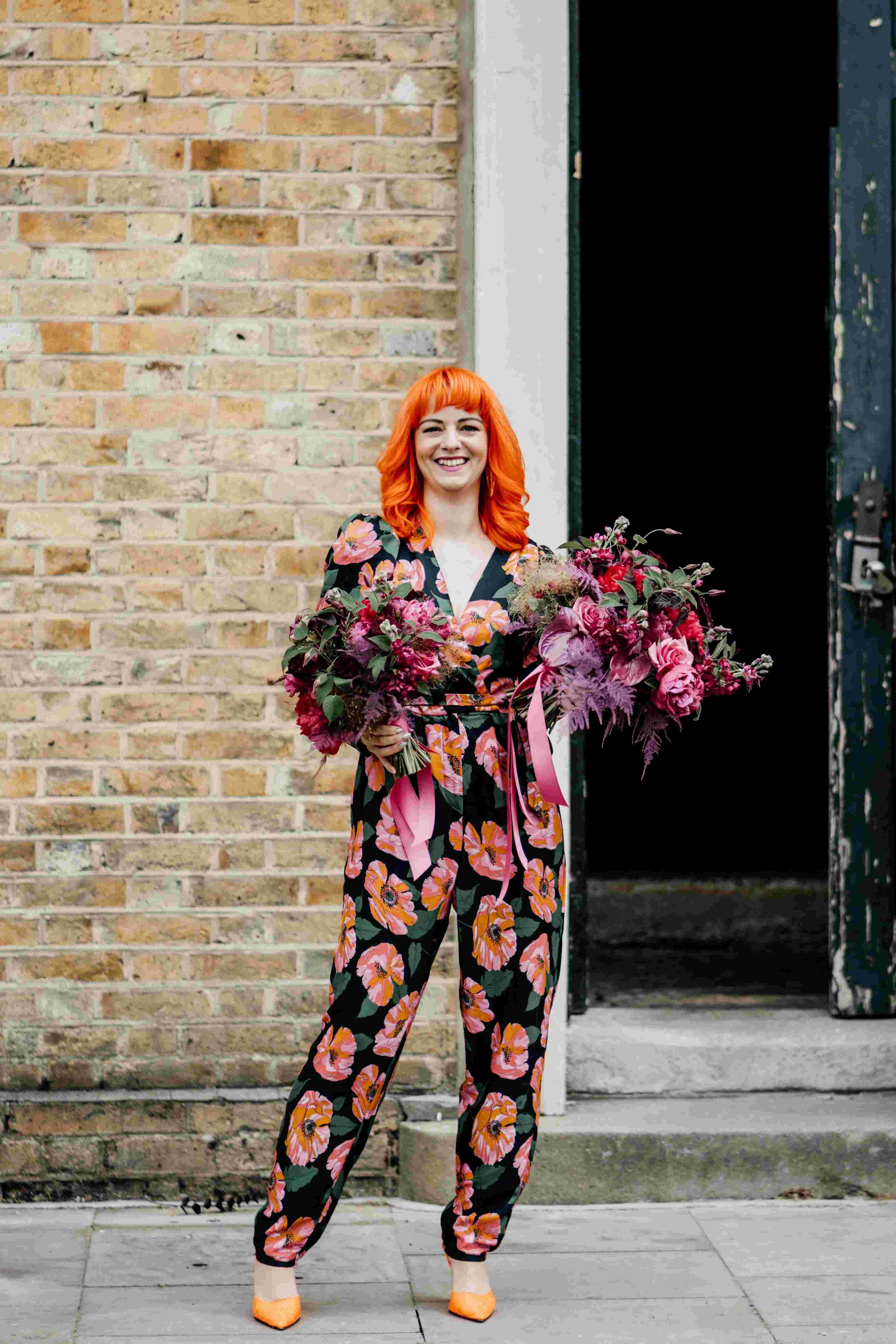  Photo by: Tom Wyatt Photography  Non-traditional humanist celebrant is stood outside, smiling at the camera and holding two large colourful wedding bouquets. She is wearing a floral long sleeved jumpsuit and orange heels which match her hair.  