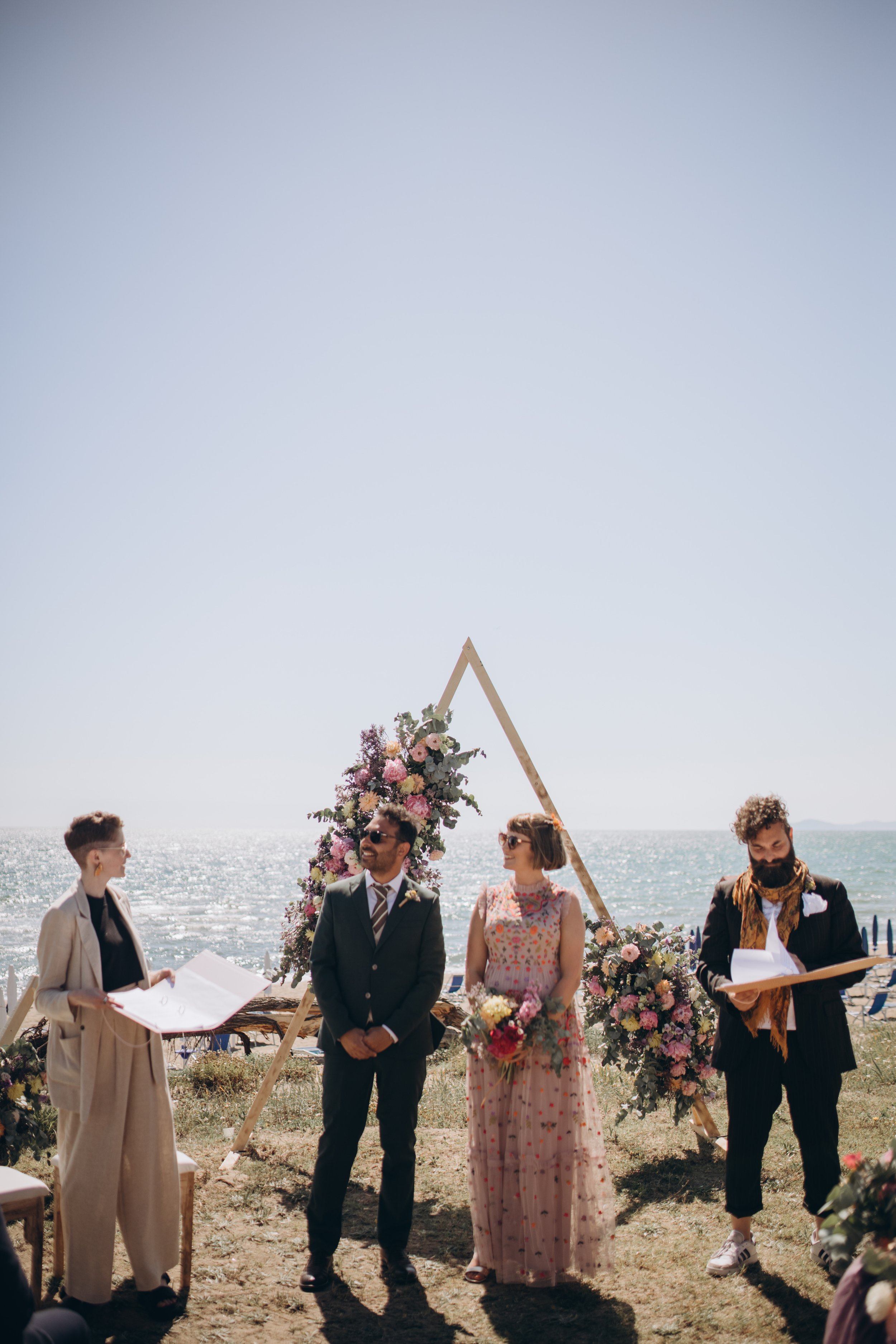  The cool, modern celebrant is stood on a beach next to the bride and groom leading their wedding ceremony as their wedding guests look on.   Photo by: Lisa Elisa Photography   