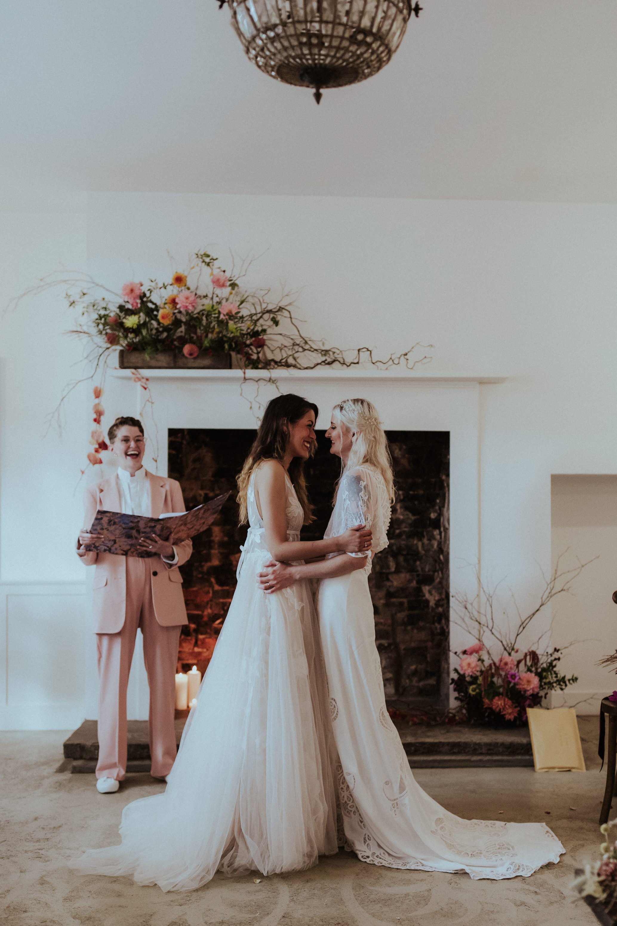  The celebrant is stood to the side of the brides with her folder open smiling. She is looking over at the brides, who are standing face to face looking into each others eyes and laughing at a moment created for their ceremony by the funny wedding ce