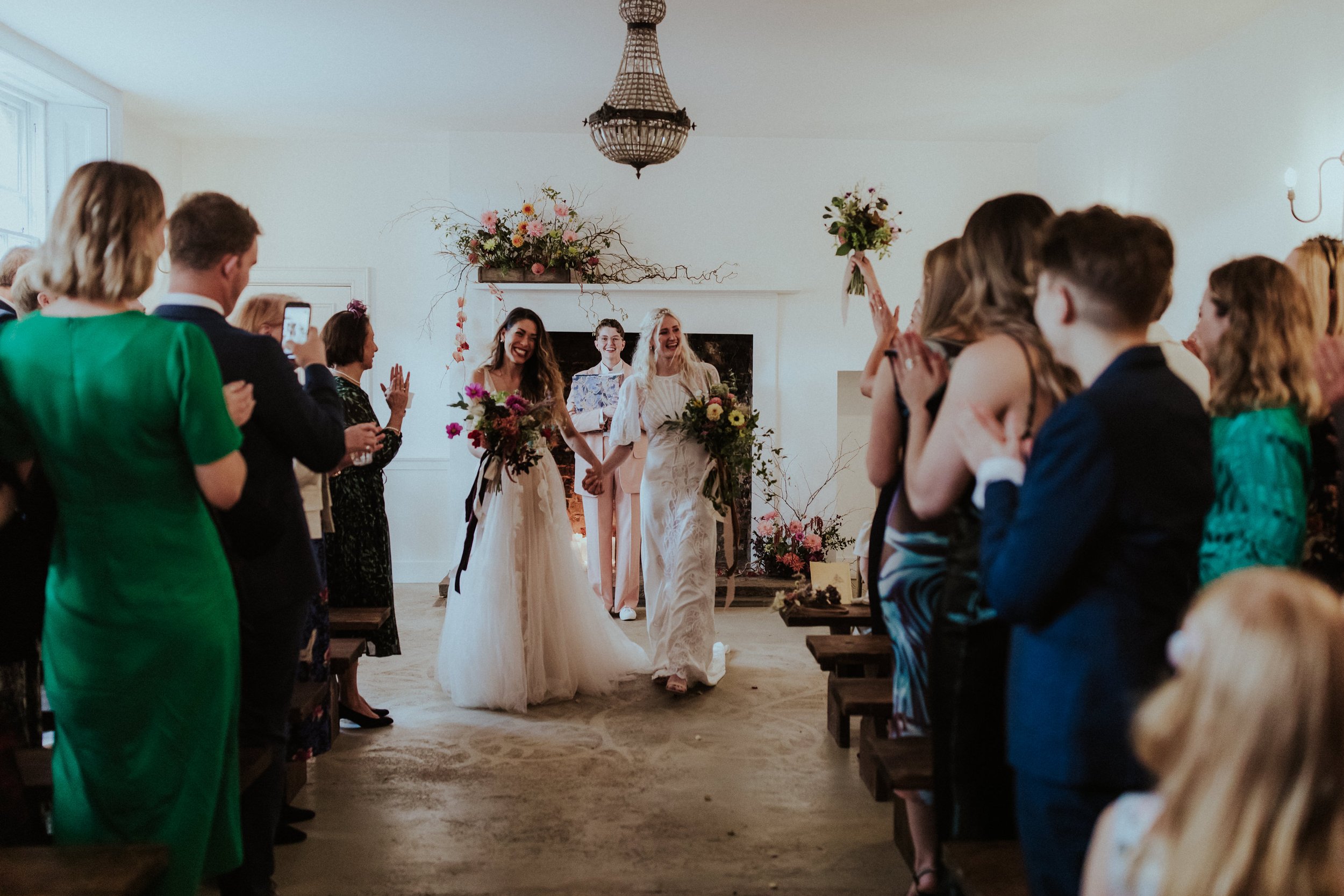  The modern, inclusive celebrant is captured stood behind the brides, smiling after delivering a fun and warm hearted wedding ceremony for the couple.  The smiling newlyweds are holding hands as they walk down the aisle past their wedding guests.  Ph