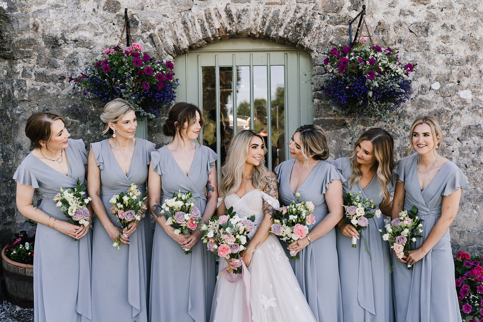  The bridal party are all stood holding their romantic, pastel wedding bouquets in front of them.  The bride is standing in the middle of her bridesmaids who are all looking towards her.   Photo by Oliver Rees Photography 