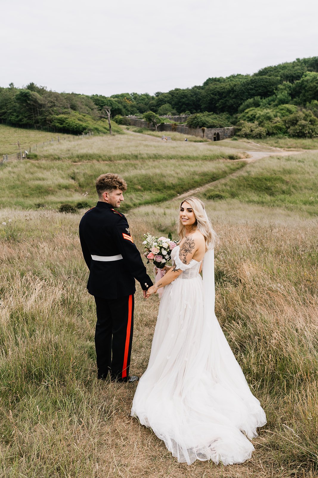  Bride and groom are walking across a field hand in hand. The bride has turned back towards the photographer and is holding her whimsical, romantic bridal hand tied bouquet made up of soft pink and cream florals.  Photo by Oliver Rees Photography 