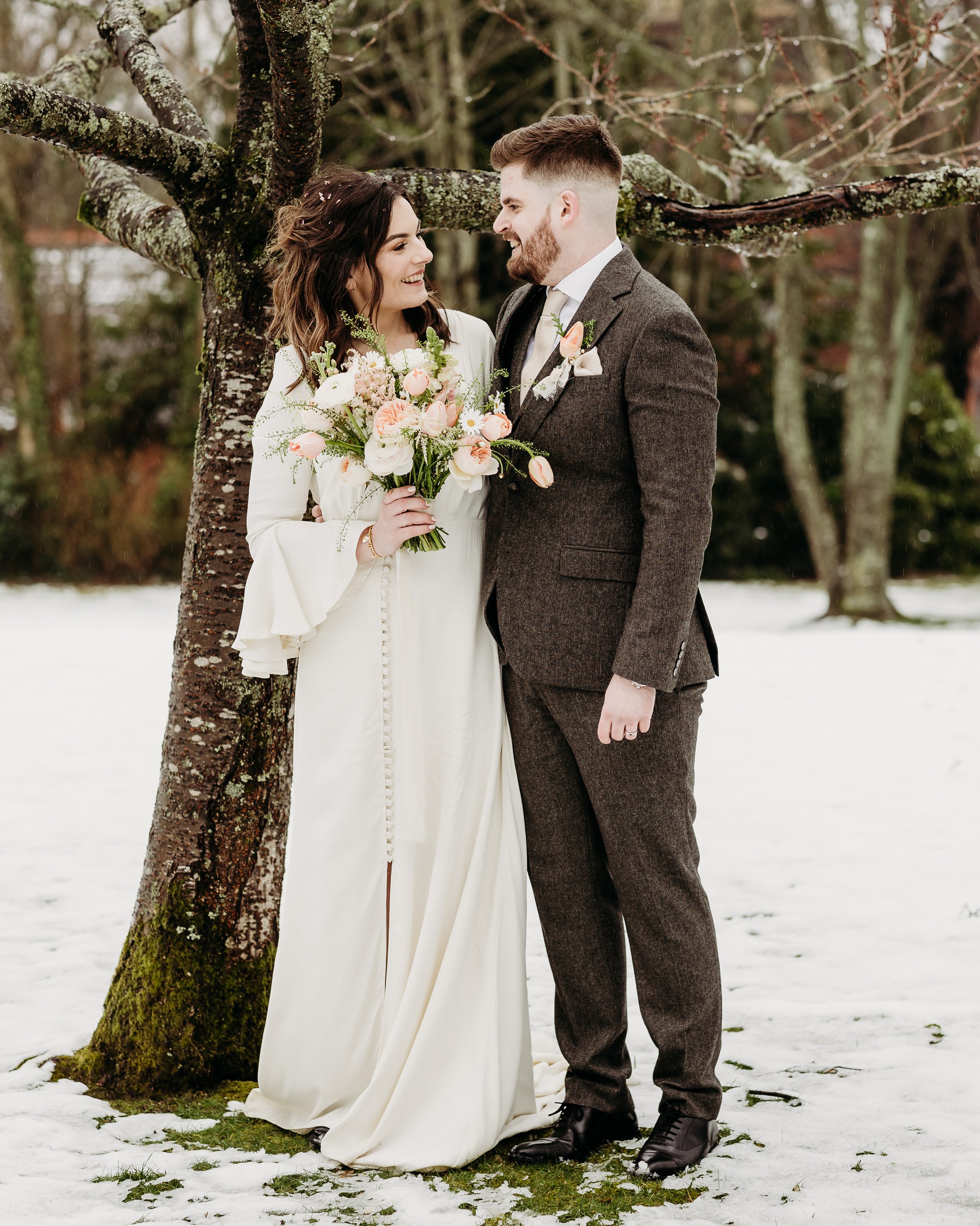 The bride and groom stood on snowy grass next to a tree trunk looking into each others eyes. The wild, hand tied bridal bouquet is both romantic and striking in its softness.    Photo by: When Charlie Met Hannah Photography 