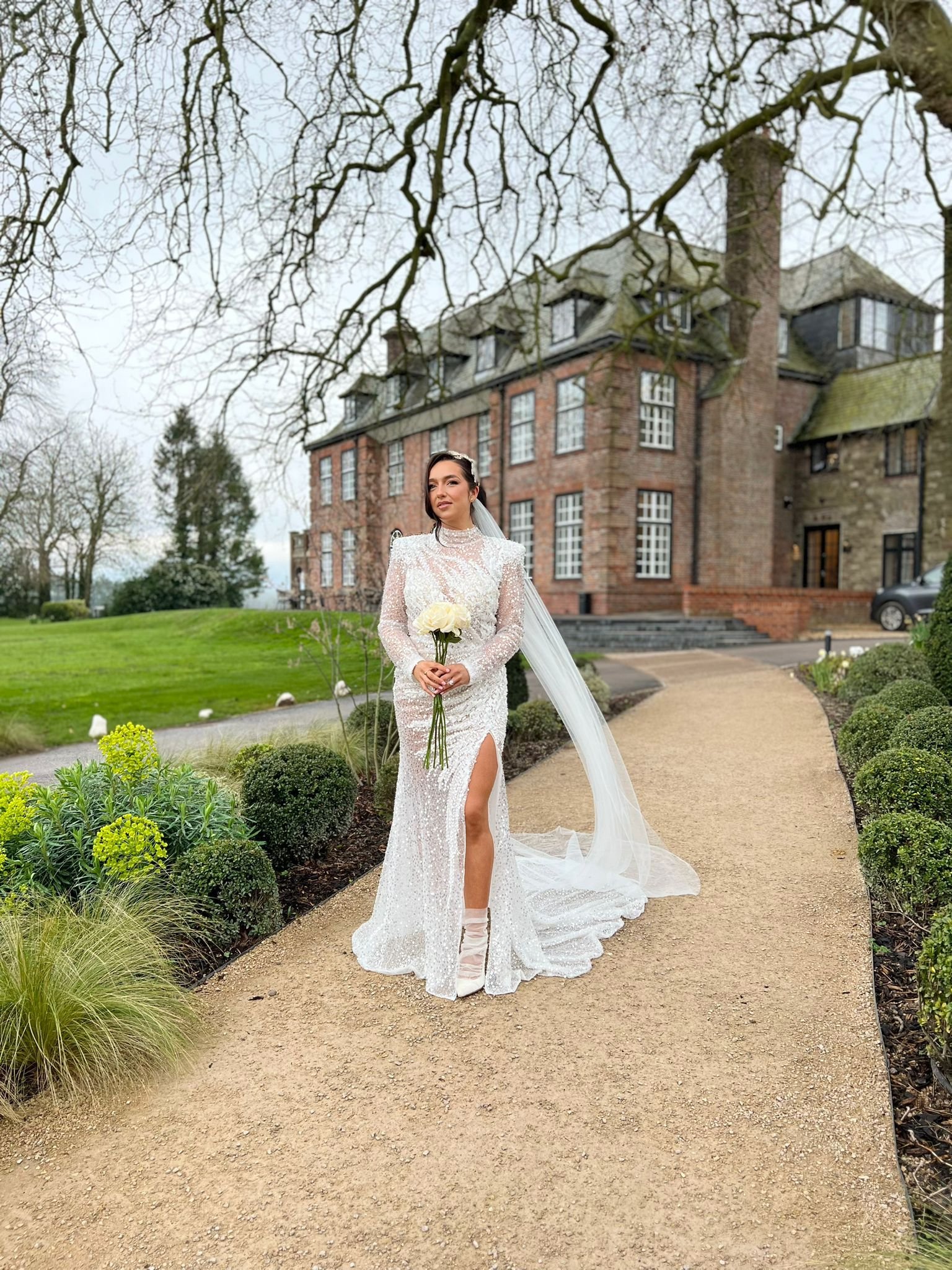  The bride is stood outside in the grounds in front of a grand building. The modern bride is wearing a long, sparkly white wedding dress with long sleeves and a thigh high split at the front along with white ankle boots. The sleek design of the white