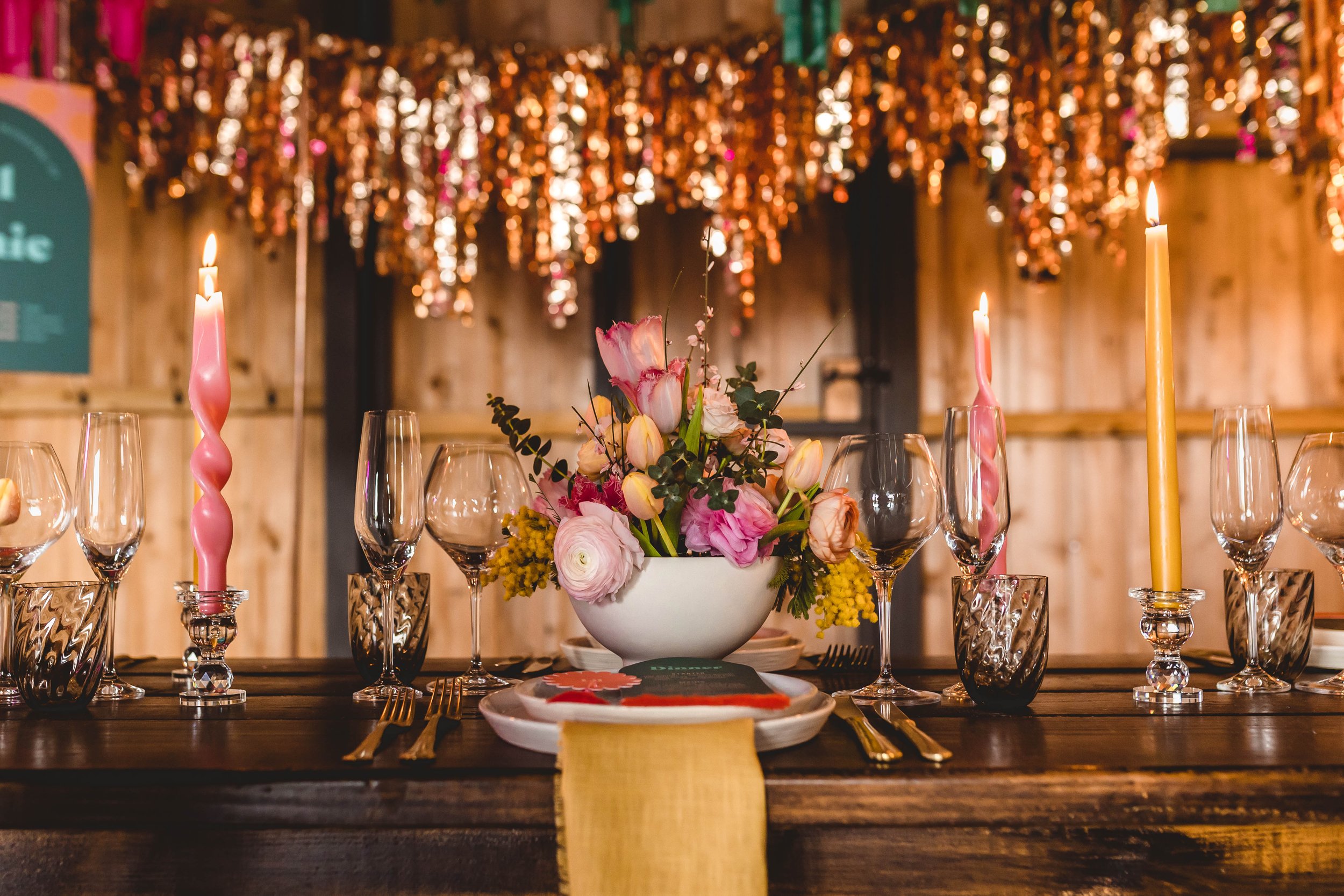  A modern display of bright, wild flowers are displayed in a white bowl in the centre of the wedding table. The softness of the pink and yellow florals contrasts against the bare wooden walls of the room and the gold streamers.  Photo by: Carmel Tere