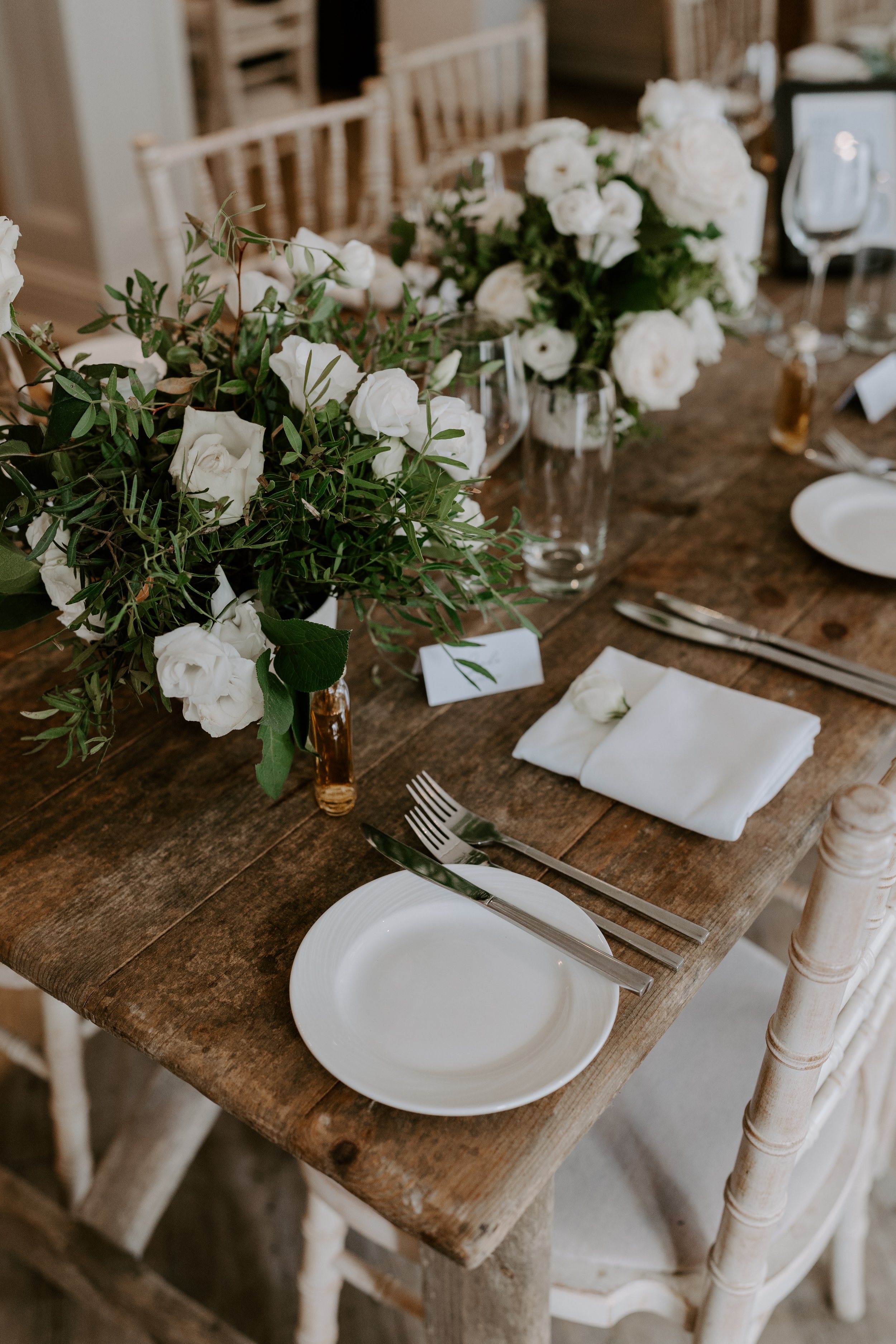  A wooden wedding breakfast table looks stylish with white crockery and modern floral arrangements down the middle made up of of white roses and green foliage.  Photo by: Marta D Photography @cmartad.weddings 