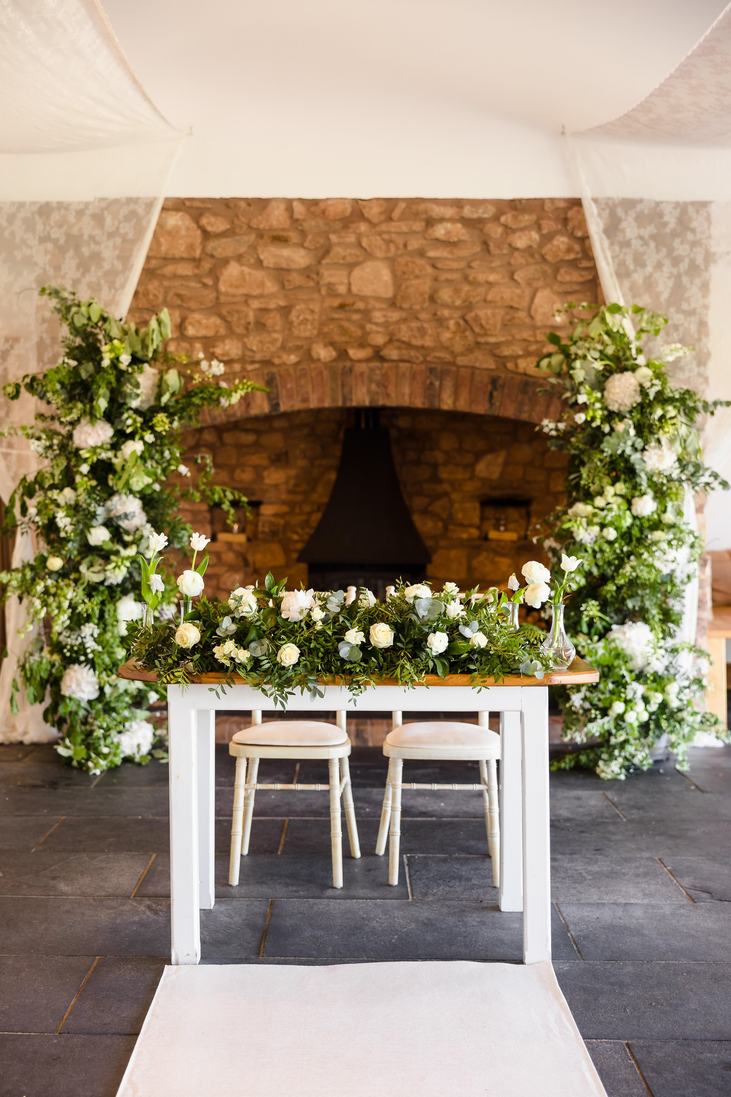  A bespoke, modern, wedding floral installation. The stylish wedding installation is placed either side of a brick fireplace and uses large headed white florals and greenery. The same flowers and foliage are used across the front of the top table lai
