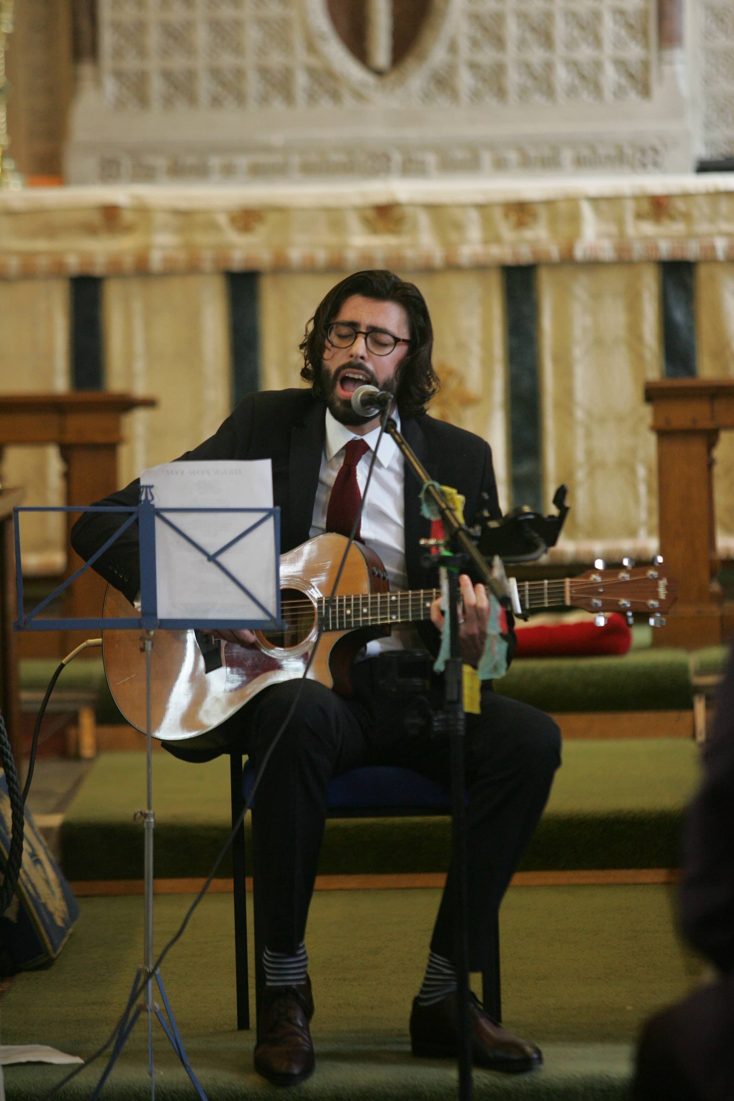  An image of the acoustic musician sat down with his acoustic guitar performing one of his indie-style folk songs as part of the wedding ceremony. 