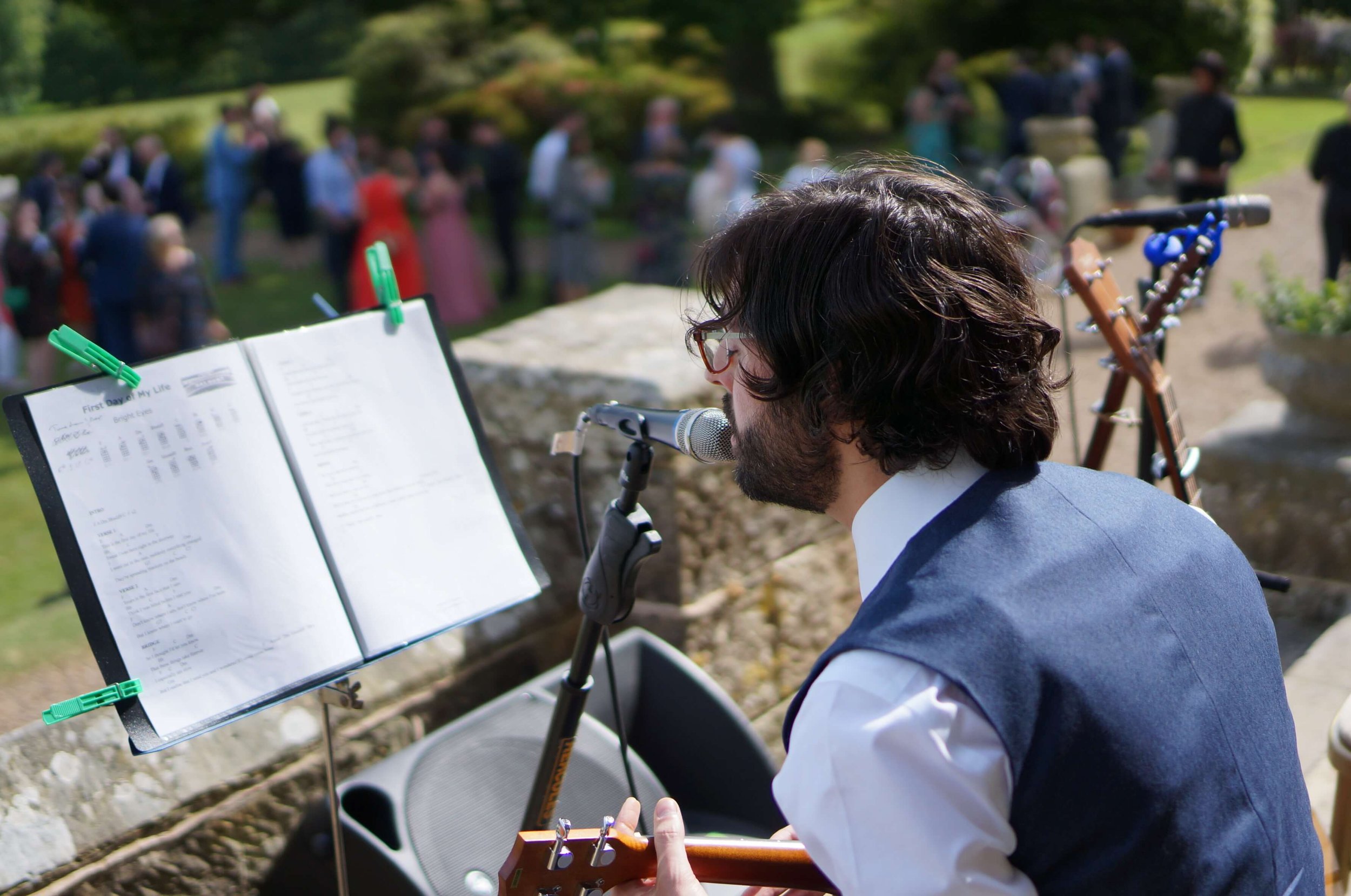  The indie-folk acoustic musician is sat outside in the sunshine entertaining a crowd of wedding guests as he plays his acoustic guitar and sings. 