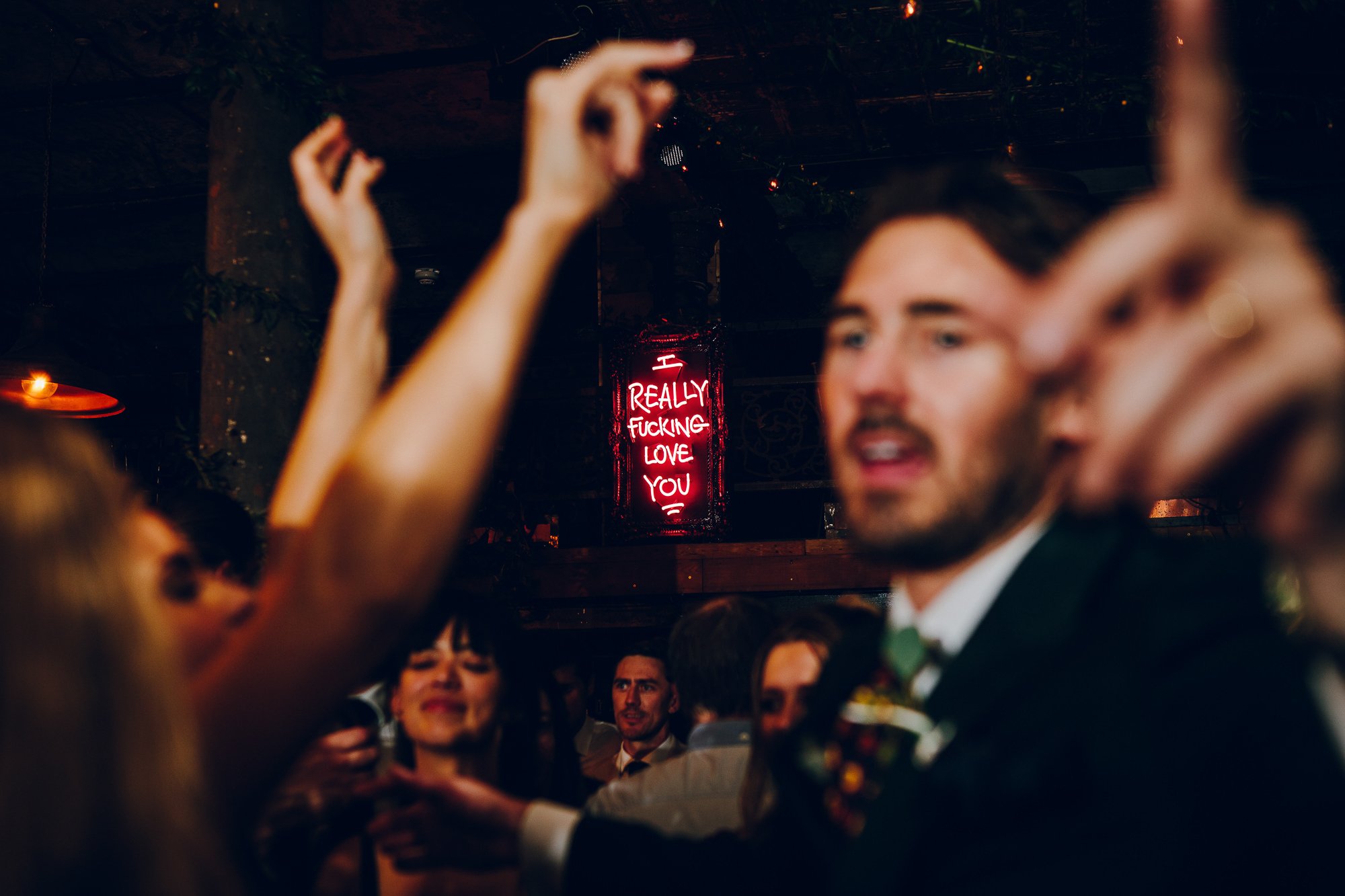  REALLY FUCKING LOVE YOU pink neon sign is clear in the background of this cool, modern image where the wedding guests are pictured blurred as they dance.  