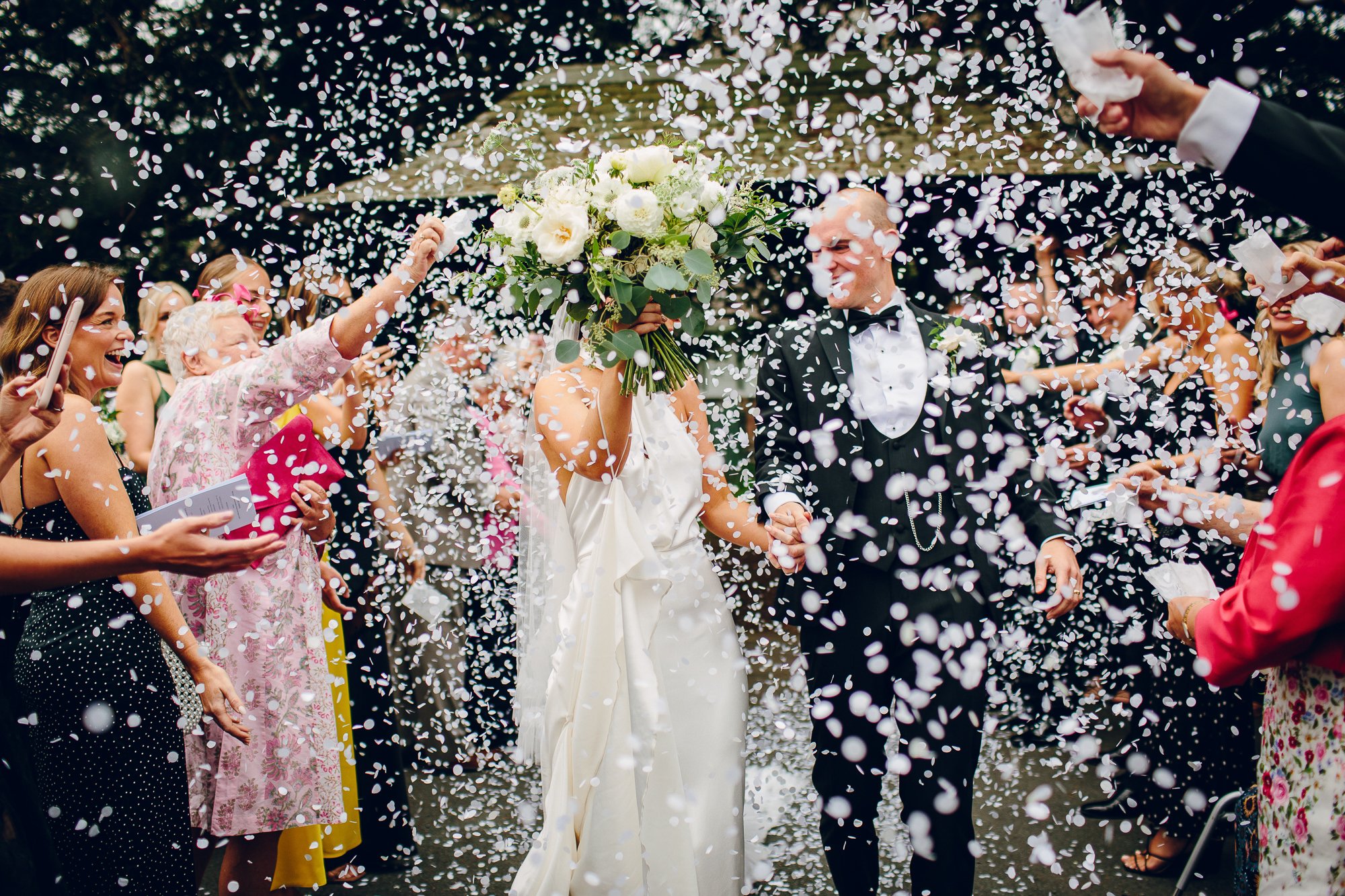  Stunning creative shot captured by the wedding photographer. The bride is holding up her floral hand tied bouquet in front of her face, whilst holding hands with the groom as they walk through their wedding guests who are showering them with confett