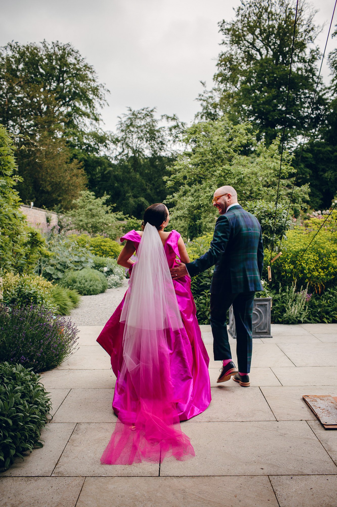  A natural moment caught on camera by the documentary style wedding photographer as the bride and groom walk away from the camera, holding hands and laughing as they make their way towards a garden in the grounds of their wedding venue. The bride is 