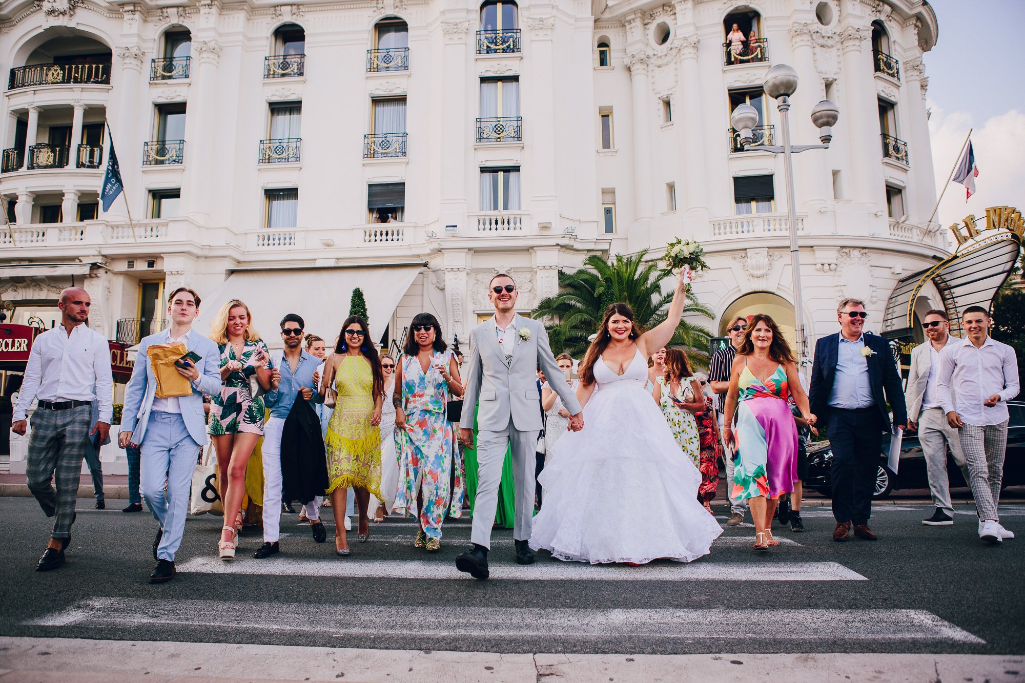  The creative, bright style of the wedding photographer is showcased in this image. The bride and groom are walking across a zebra crossing with their wedding guests walking behind and to the side of them, all wearing colourful, summery outfits. 