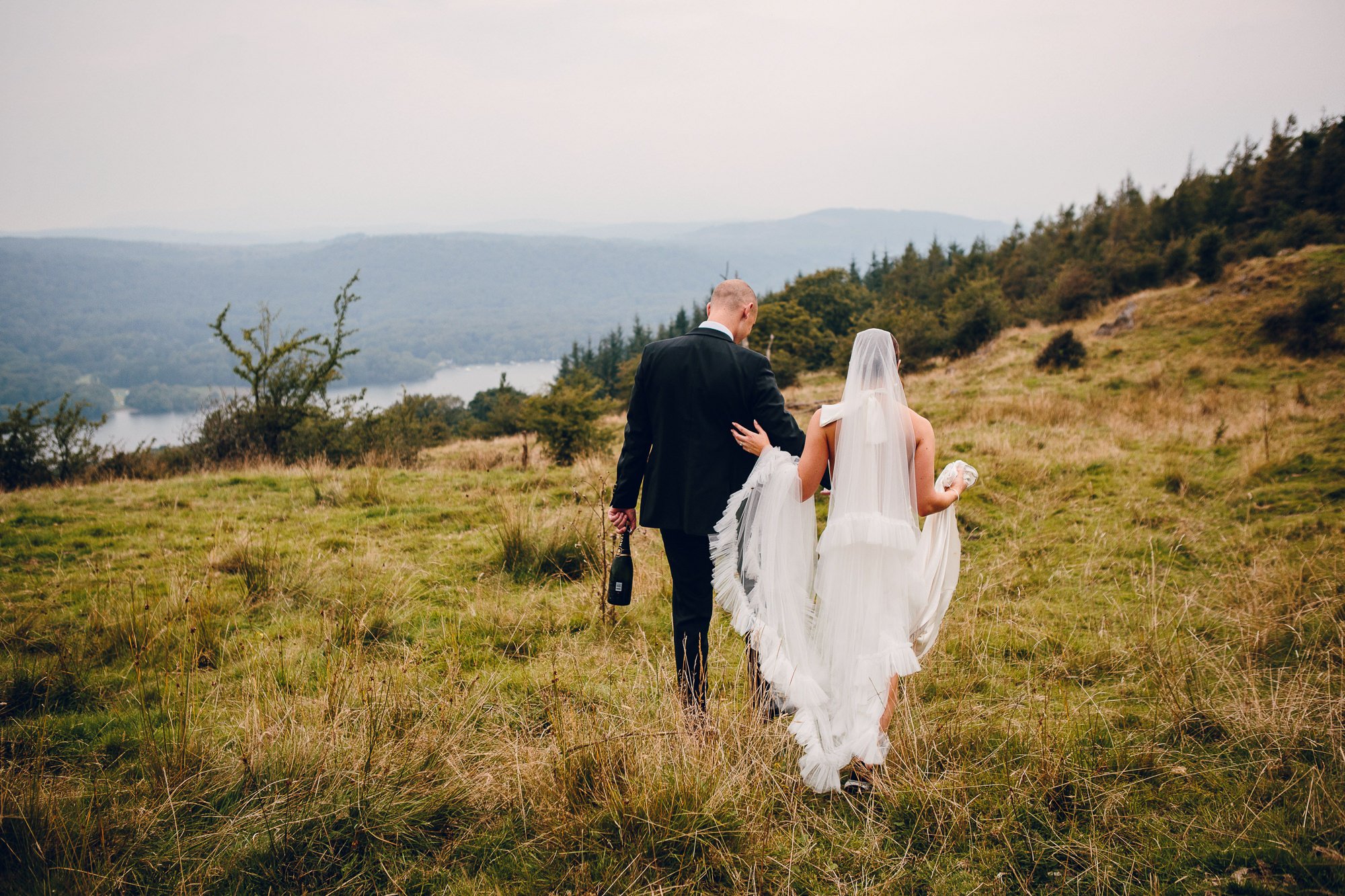  A beautiful moment captured by the creative, documentary style wedding photographer of the bride and groom as they have a moment walking together across a grassy hillside. The bride is holding onto the grooms arm and has her dress lifted up over her