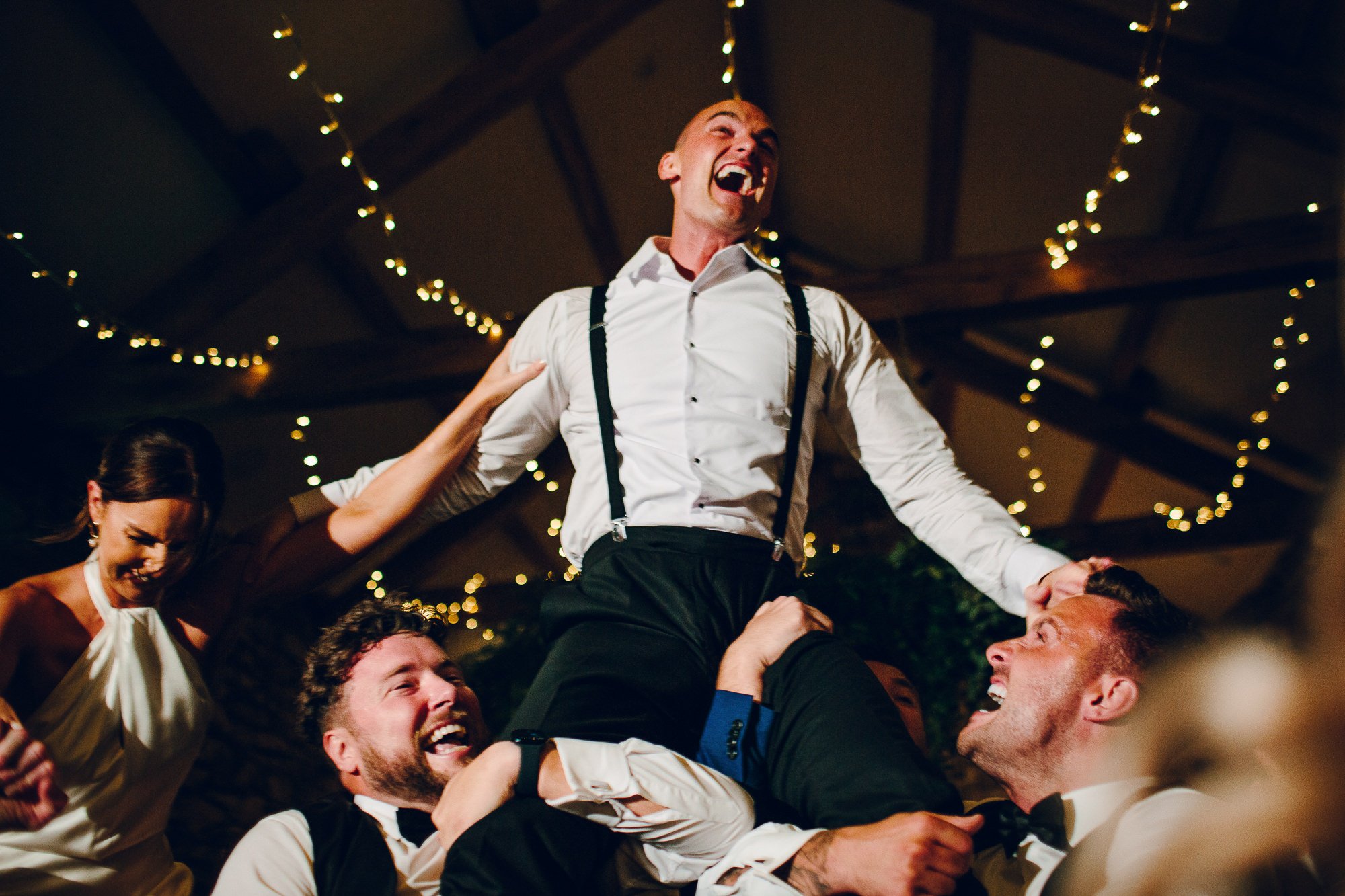  The wedding photographer has captured the moment when the groomsmen lifted the groom up onto their shoulders during the wedding reception. They are all laughing and smiling as they dance. 