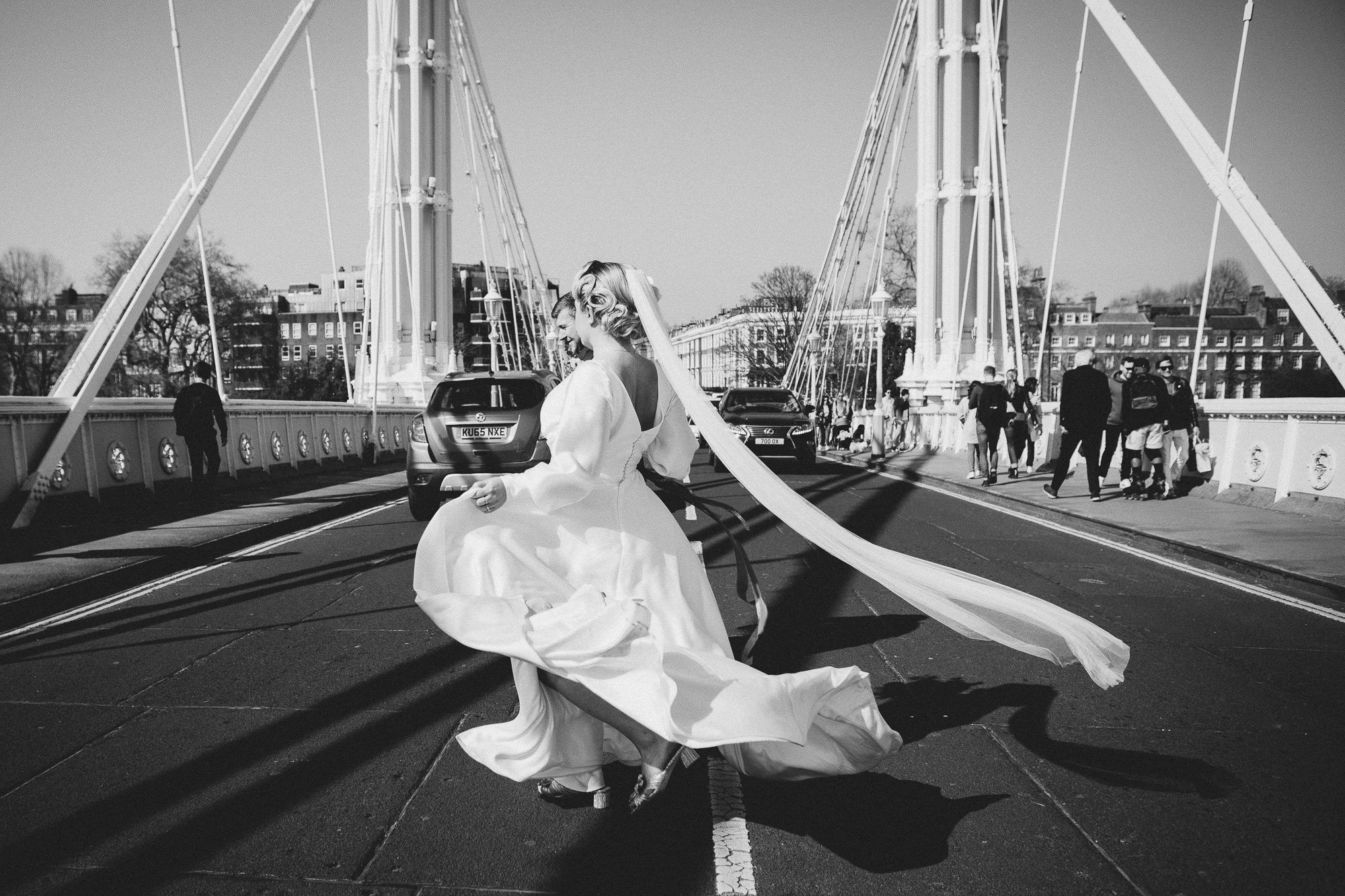  A cinematic style black and white image of a natural moment caught by the wedding photographer as the bride and groom cross the road going over a suspension bridge.  