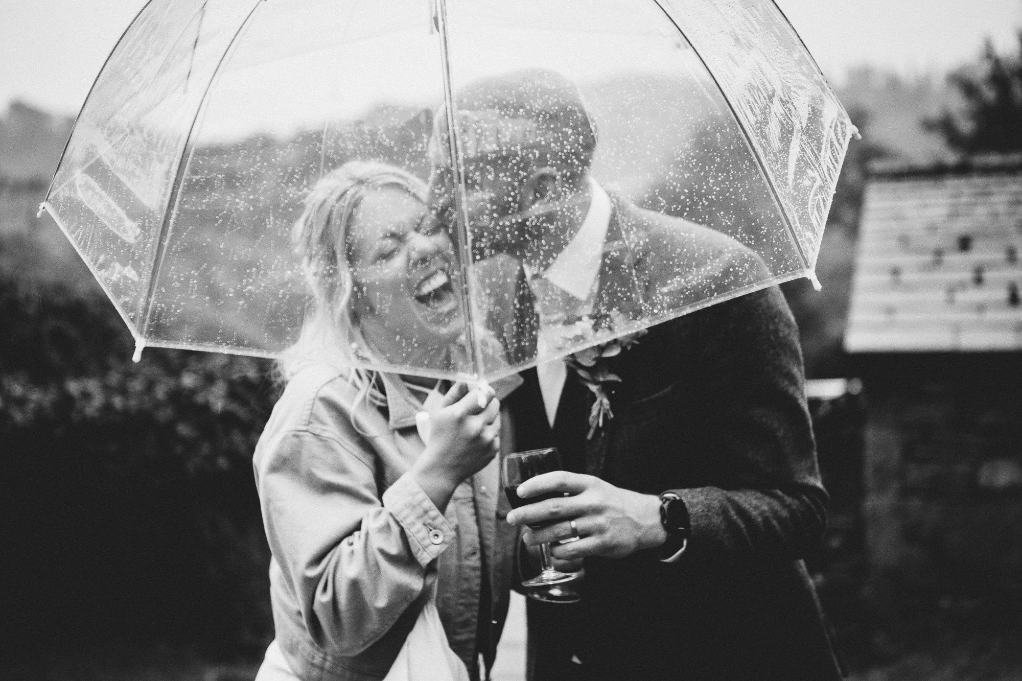  A black and white photo of a beautiful moment documented by the wedding photographer. The groom is kissing the brides head as she is stood laughing next to him, whilst they are under a see-through umbrella in the rain. 