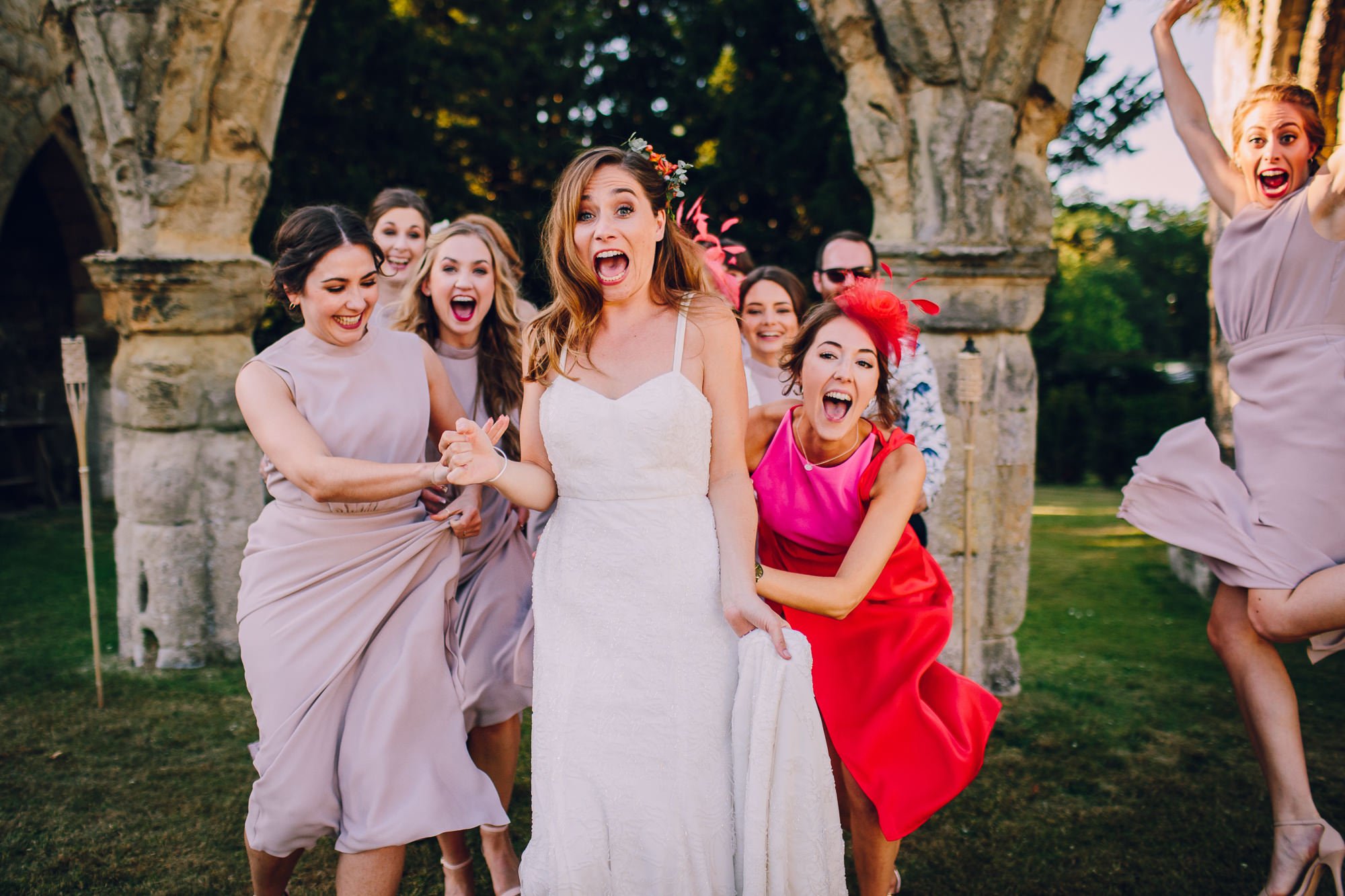  A wonderful natural moment has been captured by the wedding photographer in this bright, fun image. The bride is stood with her mouth open in surprise, outside with ruins in the background behind her. The bridesmaids are in a group behind the bride 