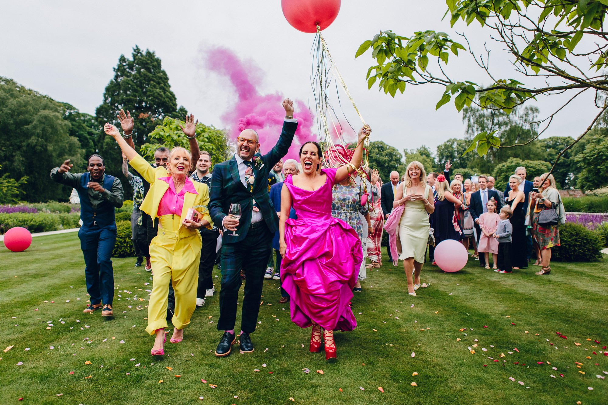  A colourful image taken by the wedding photographer. The creative composition captures the bride and groom leading the wedding party across the grass. The bride is wearing a bright pink wedding dress and carrying a big, round, red balloon. She is wa
