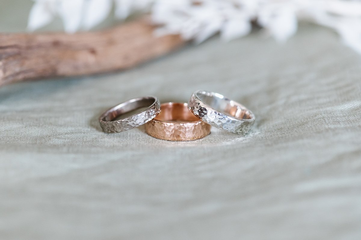  Sustainable Recycled Metal Wedding Rings. A hammered rose gold wedding band displayed in the middle with two hammered sterling silver bands on either side.  Photo by: Fiona Kelly 