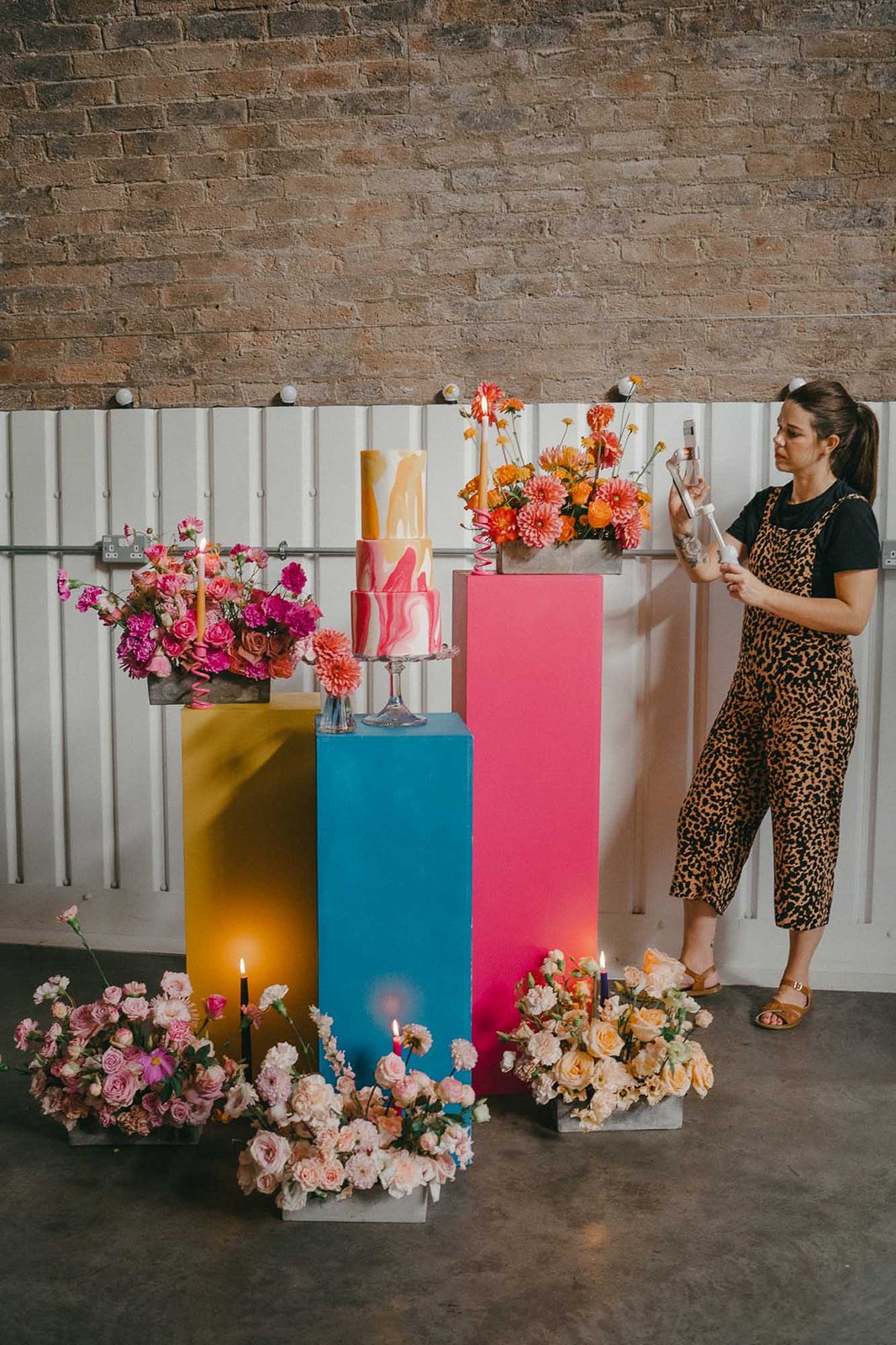 A behind the scenes moment captured by wedding content creator of florals and wedding cake being set up and displayed on bright plinths.   Photo by: Josephine Elvis 