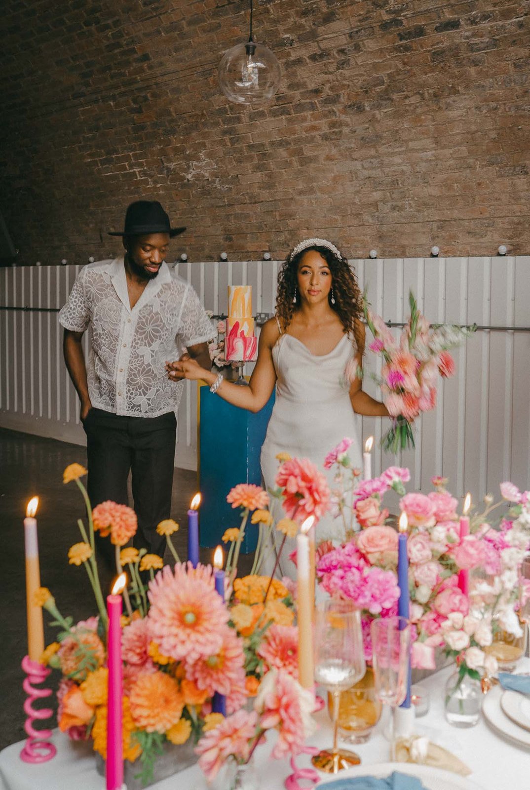  Bright florals and table candles adorn a modern wedding table as the bride and groom are walking behind it. The newlyweds have their images brought to life by wedding content creator who captures all the behind the scenes moments.  Photo by: Josephi