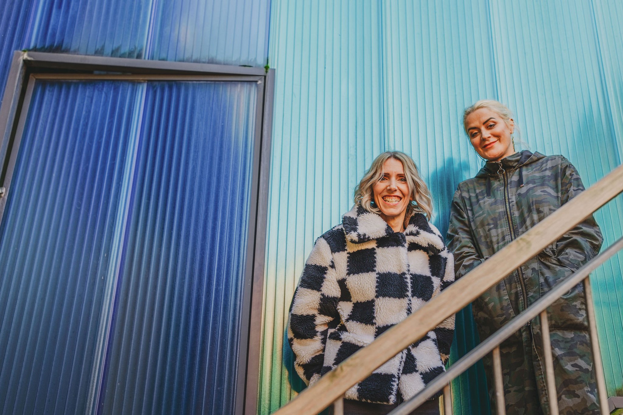  Melissa, founder of The Un-Wedding and Helen stood on metal staircase in front of corrugated metal painted blue whilst at The Un-Wedding Show Bristol. 