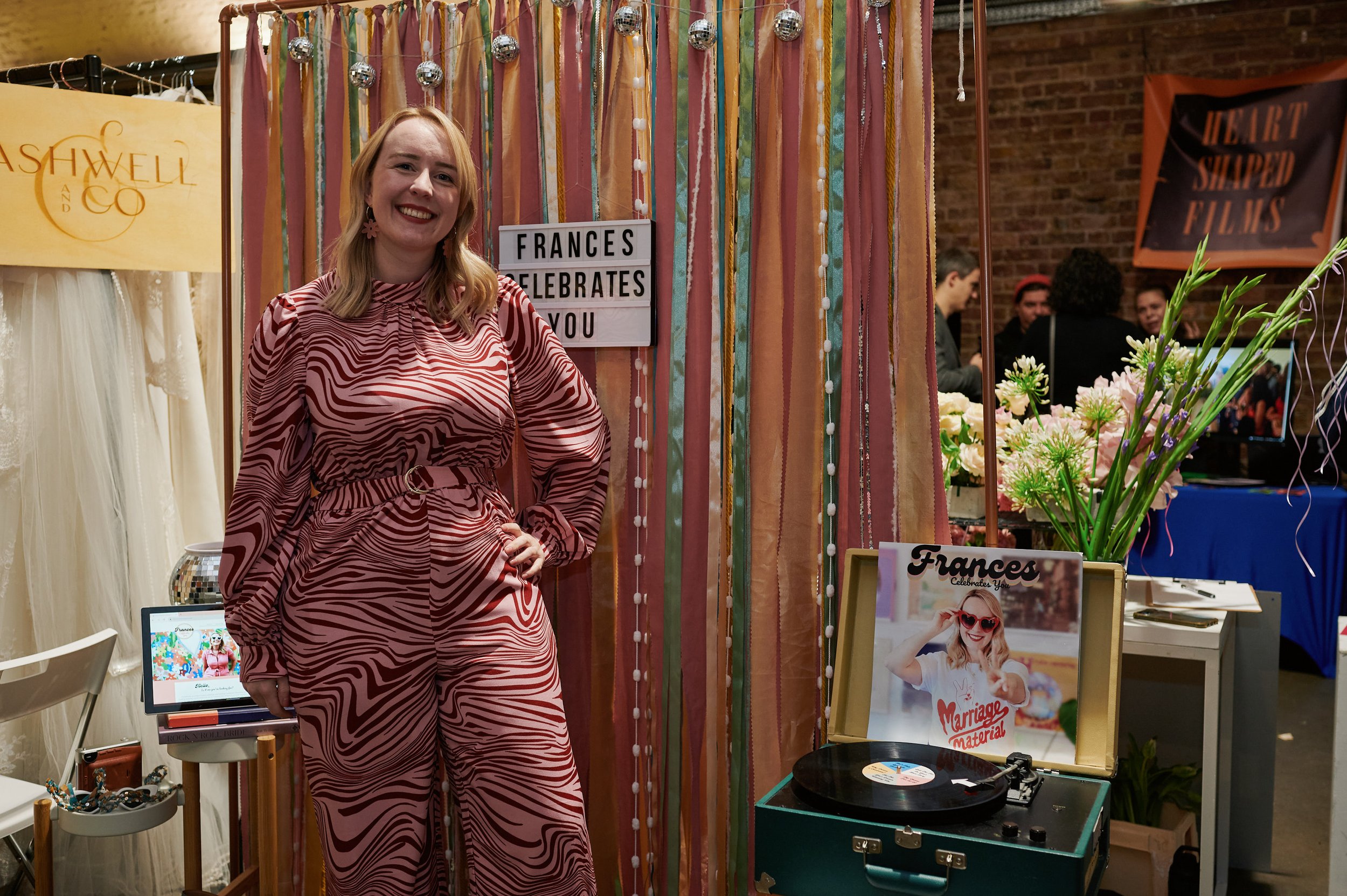  A celebrant in a pink jumpsuit standing in front of her stand at The Un-Wedding Show. There is a ribbon backdrop behind her. 