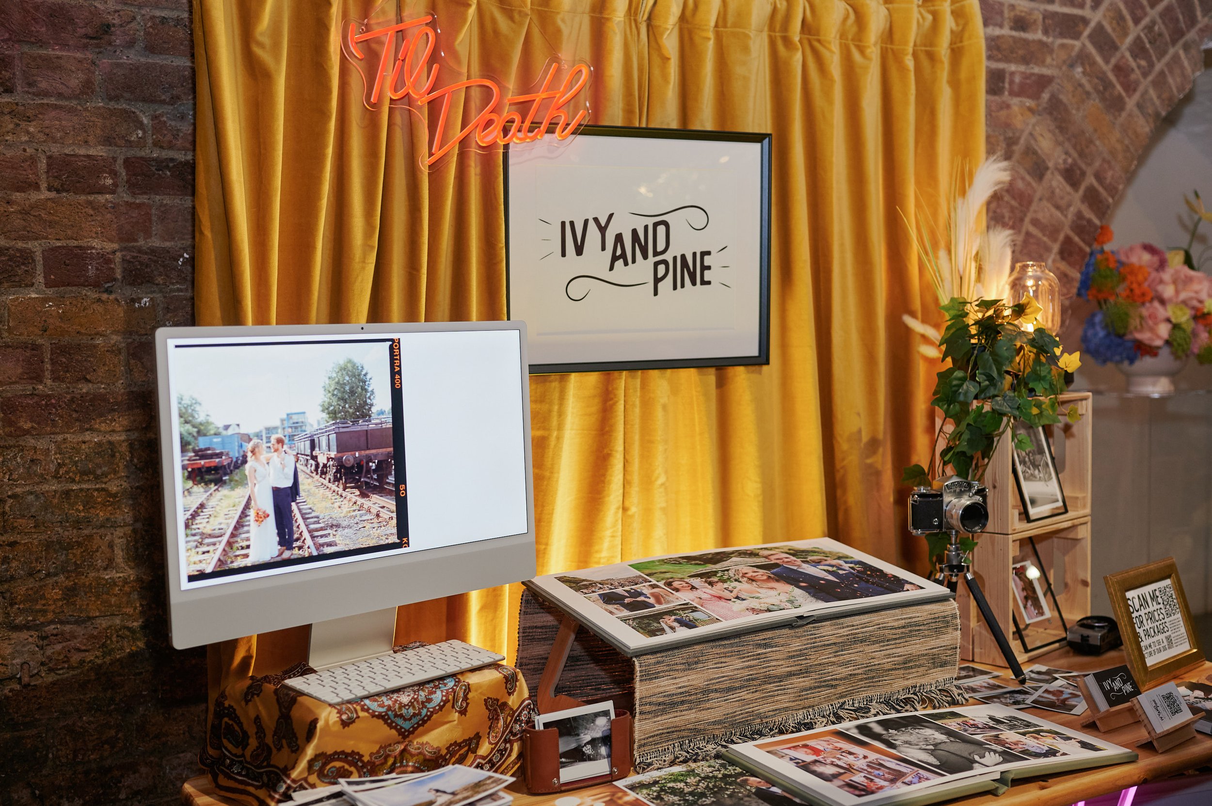  A photography stand at the Un-Wedding Show with a yellow velvet curtain with a neon sign that says Til Death. There is a sign that says Ivy and Pine and a computer displaying imagery. 