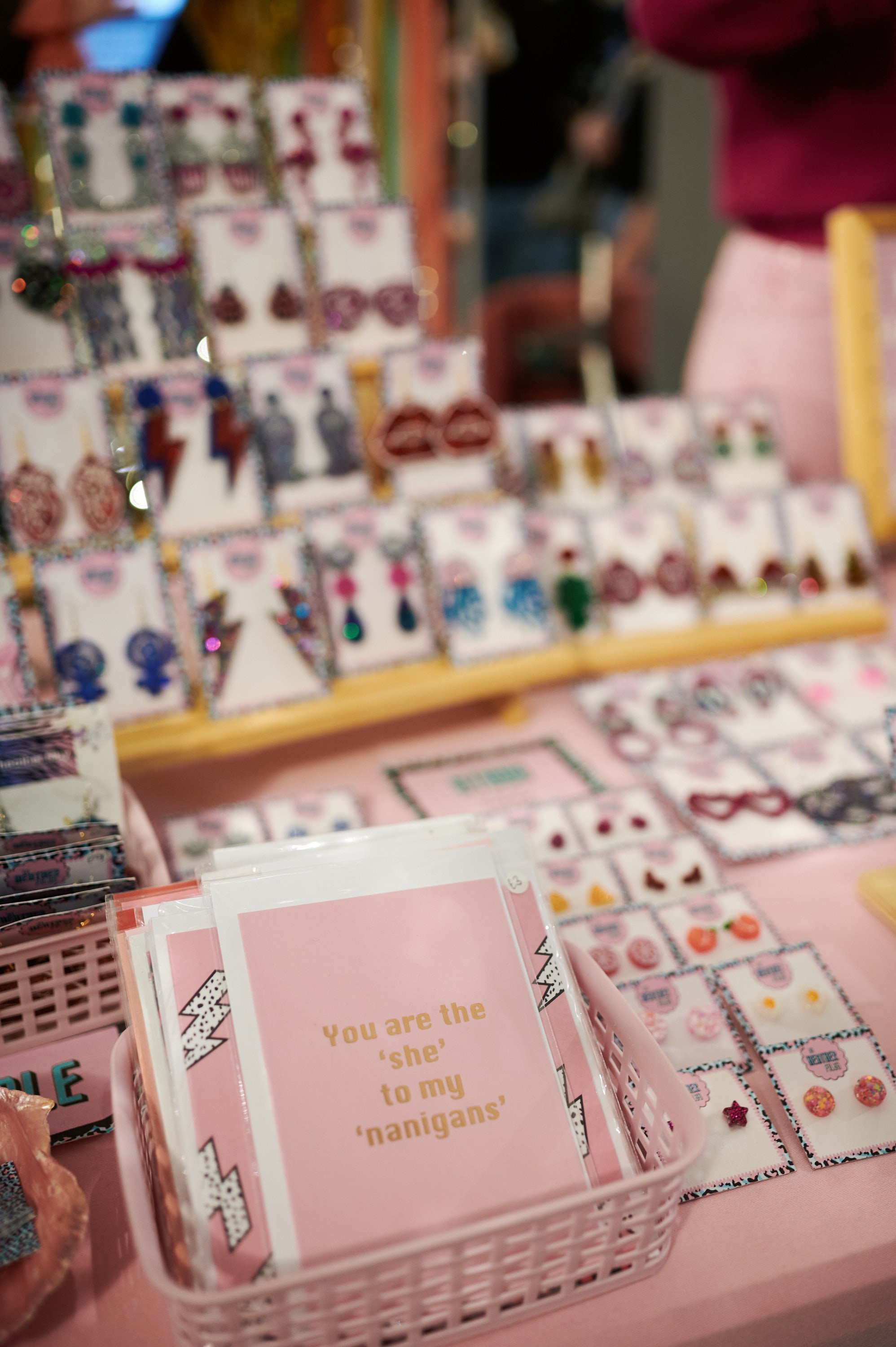  A pink table full of glittery accessories and a basket of greetings cards. One of the cards says ‘you are the ‘she’ to my nanigans.’ 