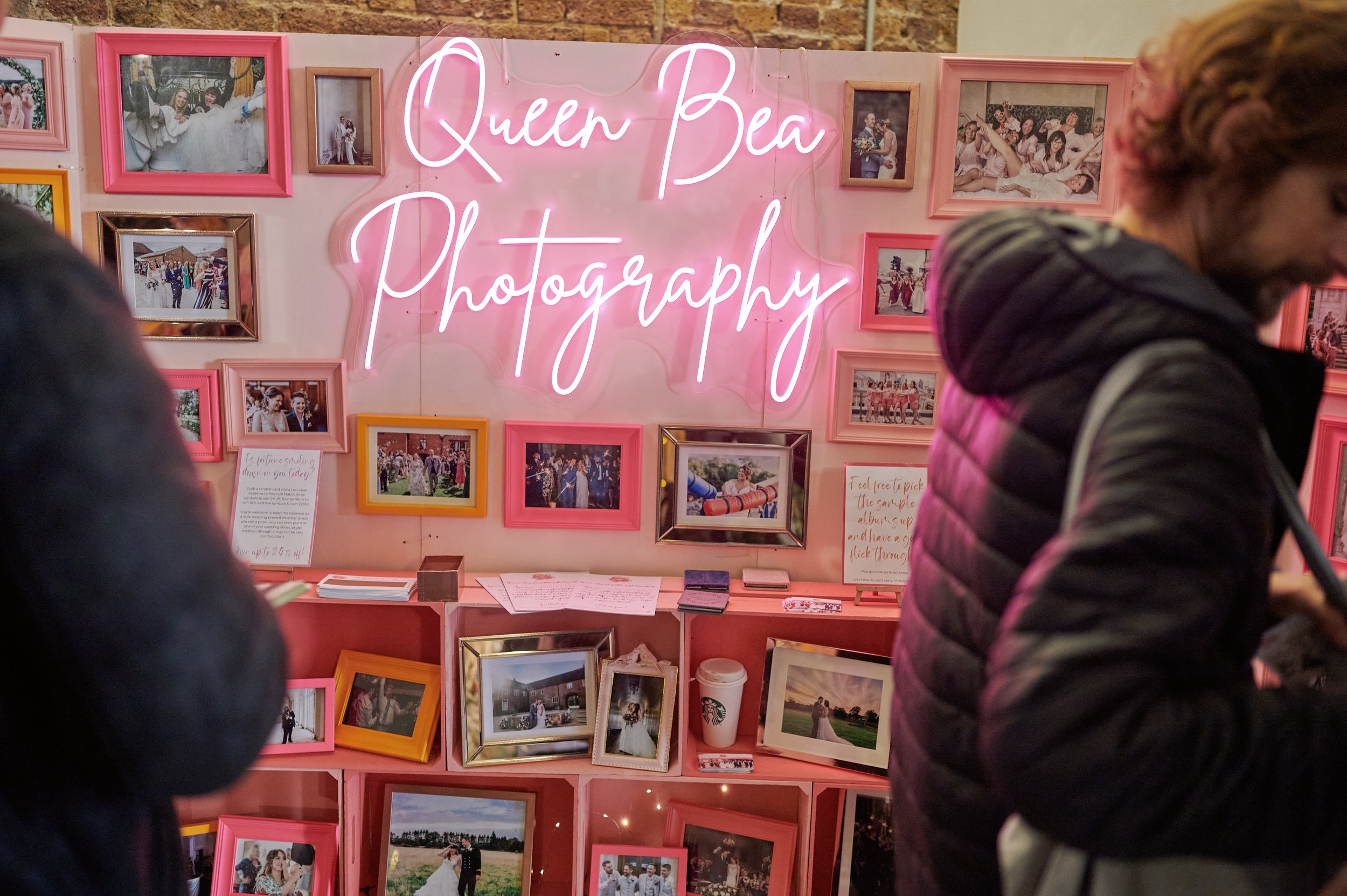  A photography stand at The Un-Wedding Show. There are lots of framed pictures and a pink neon sign that says ‘Queen Bea Photography’. 