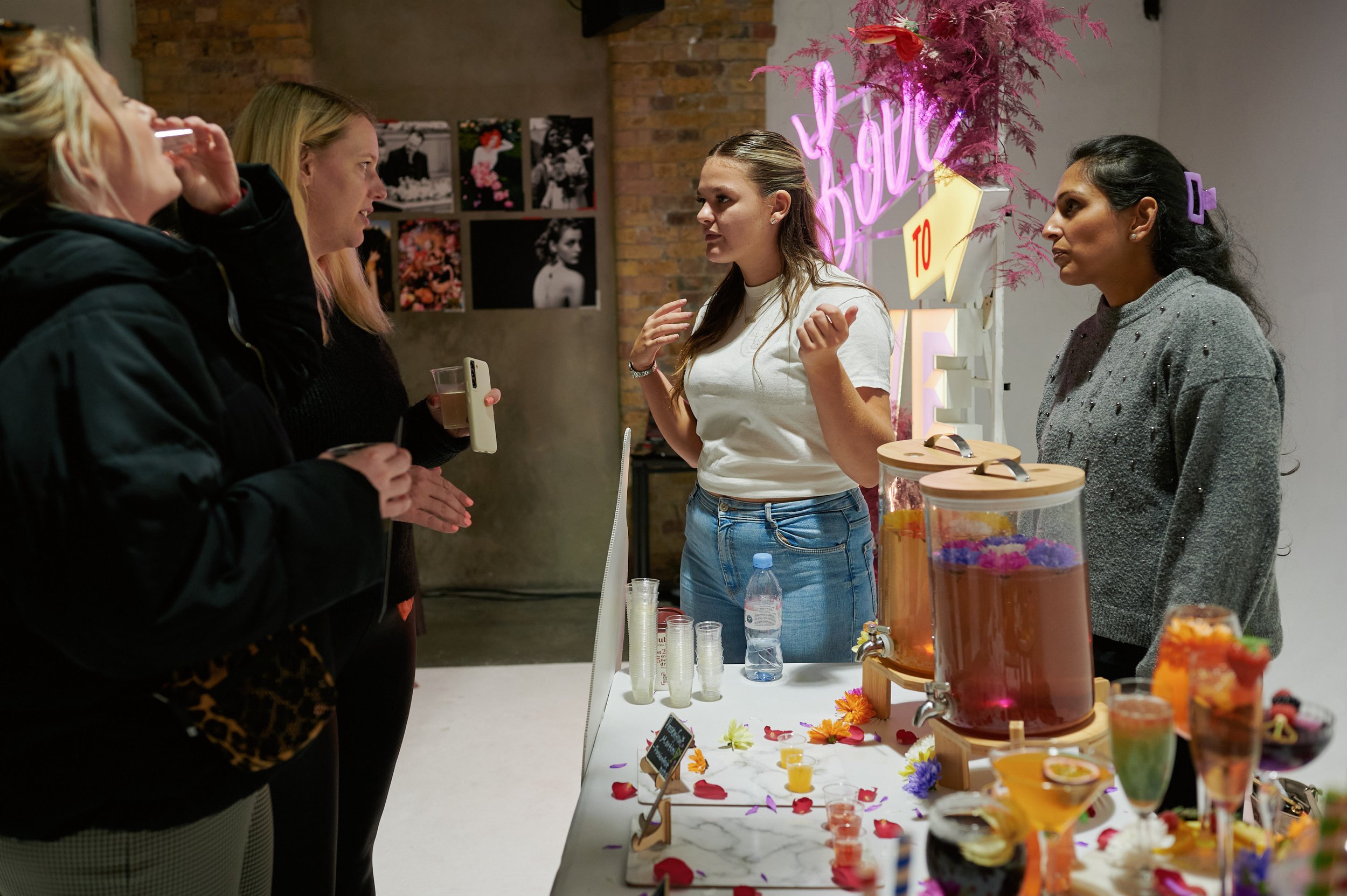  A tall woman with blonde hair is talking to suppliers at The Un-Wedding show. there is a table with some sort of floral drinks display and a neon sign in the background. 