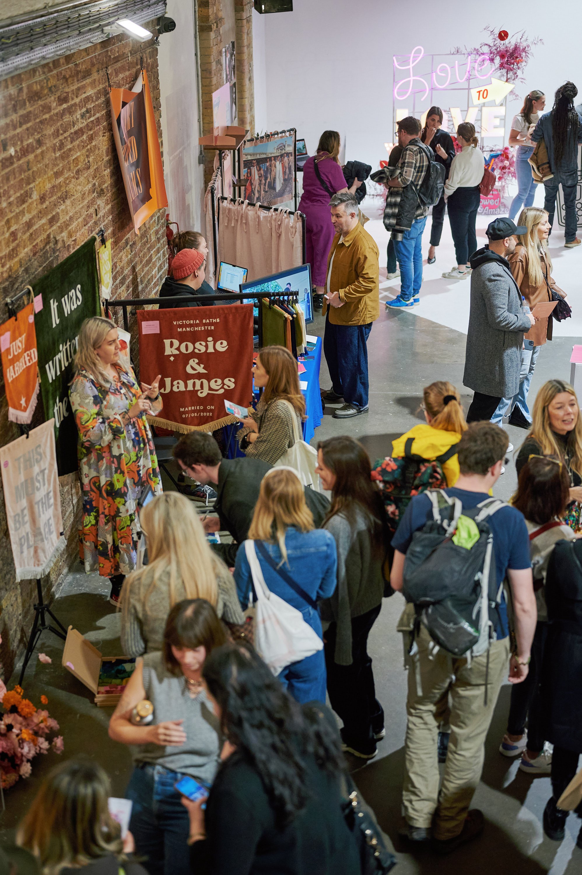  Suppliers and attendees at The Un-Wedding Show London. There is a blonde supplier in a floral dress standing amongst some colourful velvet banners. 