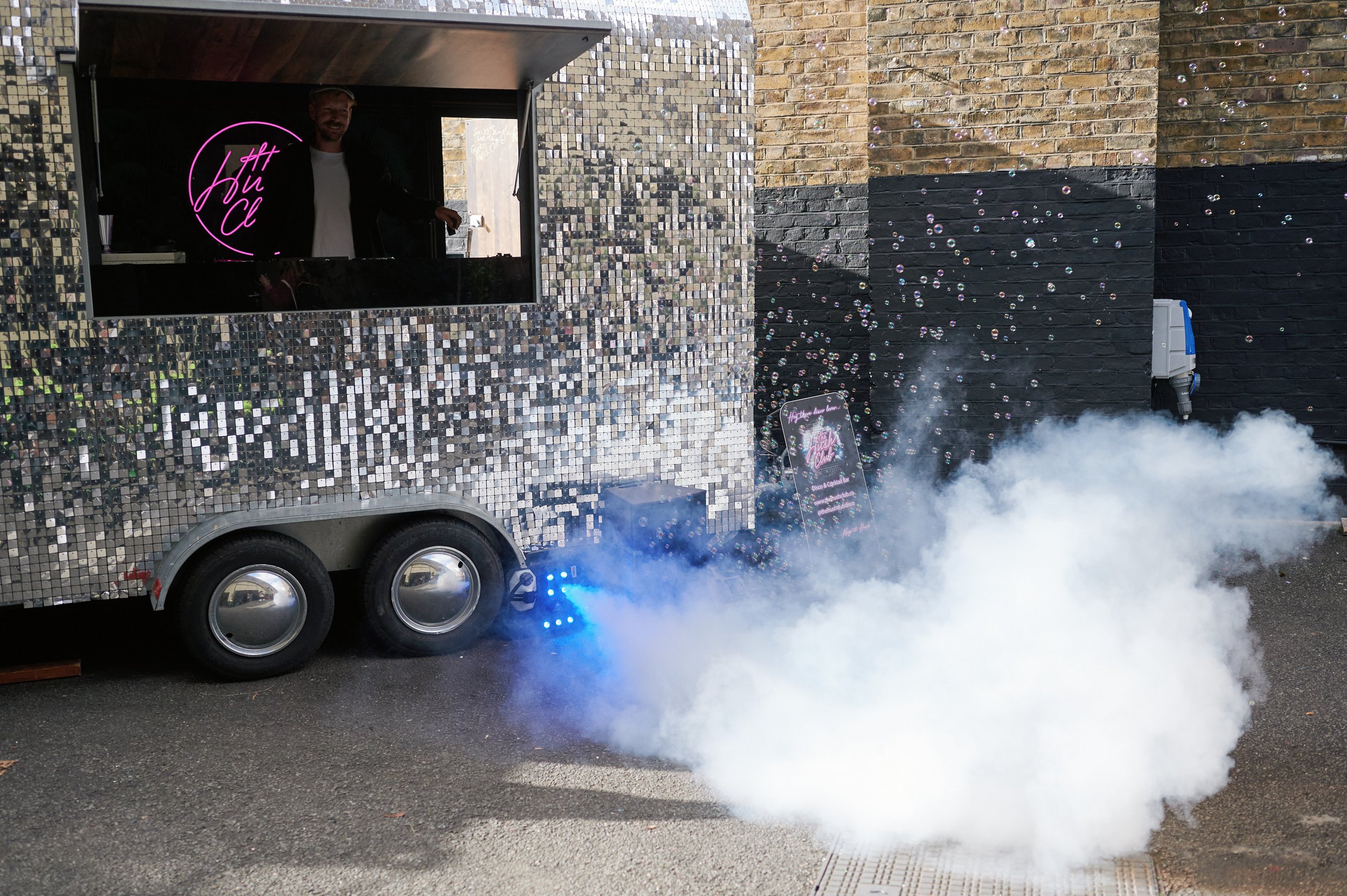 The Hush Club mobile cocktail bar. A giant silver, sequin cocktail bar, blowing smoke for effect. There is a DJ playing his tunes in there and a pink neon sign behind him that says ‘The Hush Club’. 