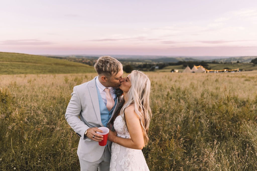  Bride and groom kissing outside. The modern wedding photographer has captured them at sunset in a cornfield with teepees in the distance behind them.  