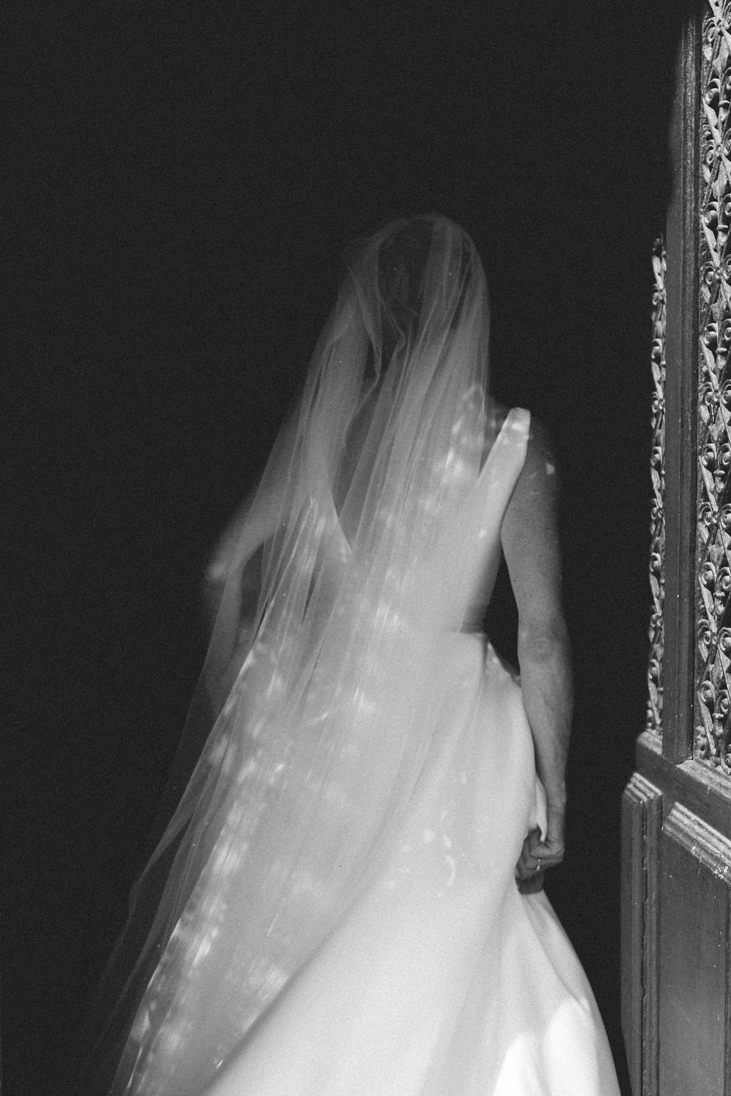  A black and white image of a bride holding her dress up slightly as she walks away from the wedding photographer. The cool image captures the beautiful natural light coming through at the side and onto the long veil trailing behind the bride over he